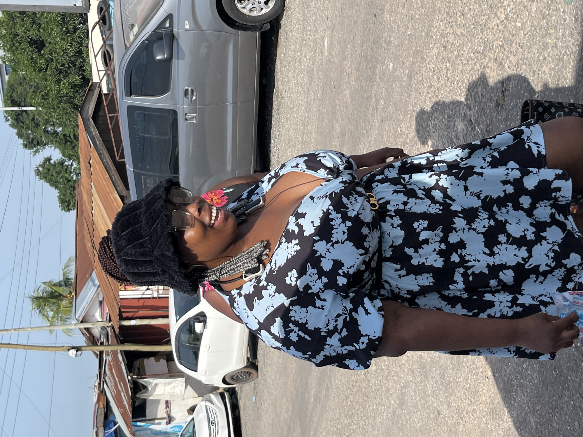 The image depicts a woman in Accra, Ghana, standing outdoors on a gravelly surface. She is wearing a black, fuzzy bucket hat, sunglasses, and a short, blue and black floral print dress with a V-neckline and a belt. Her hair is braided and appears to be tucked into her hat, with some braids extending down. She is smiling and looking to her right. The background reveals a street scene with several vehicles, including a silver minivan and two white cars. In the distance, there are informal structures with corrugated metal roofs, suggesting a commercial or residential area. Electrical wires and poles are visible overhead, along with trees and a palm tree. A person's arm, wearing a pink garment, is visible behind the woman on the left. The overall impression is an urban streetscape in daylight.