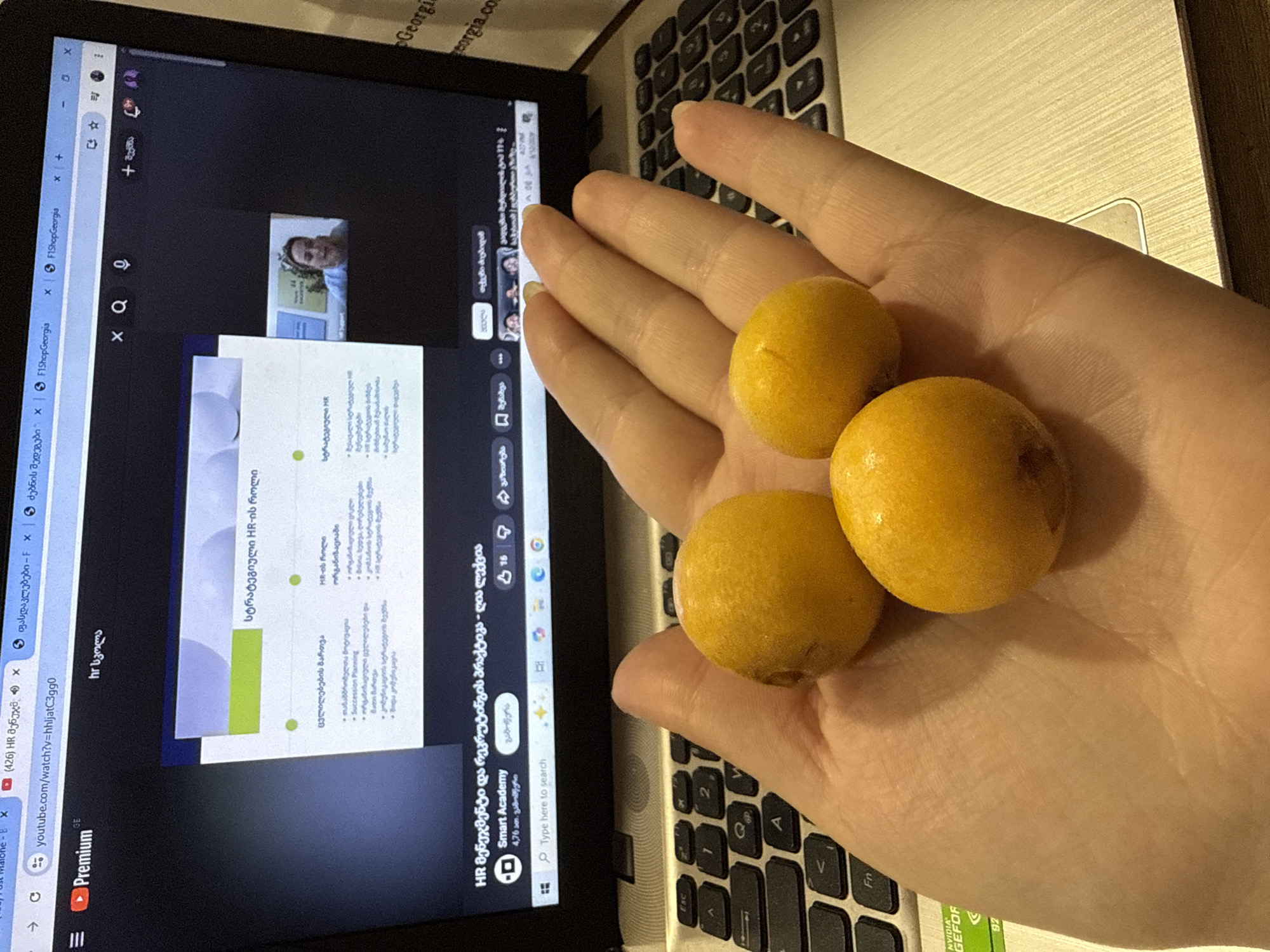 In an indoor setting, a hand holds three small, yellow, spherical fruits over a laptop. The laptop screen displays a presentation with text in Georgian, a video feed of a woman speaking, and a YouTube interface. The background shows a keyboard with visible keys like 'Esc', 'Fn', and number keys. The environment is dimly lit.