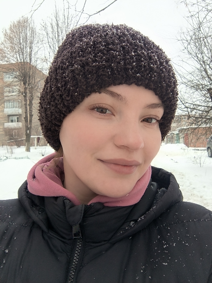A female individual is depicted in a close-up outdoor photograph taken in Vinnytsia, Ukraine, during winter. She has light skin, brown eyes, and light brown eyebrows, with a slight smile. She wears a dark brown, textured knit hat, a pink hooded sweatshirt visible at the neck, and a black puffer jacket. Small white particles, consistent with snowflakes, are present on her hat and jacket. The background shows a snow-covered urban environment. To the left, a multi-story, light-colored residential building, consistent with Soviet-era architecture, stands among bare trees. Further right, smaller structures and a partially visible light-colored vehicle are present on a snow-covered surface. The ground is uniformly covered in snow under an overcast sky.