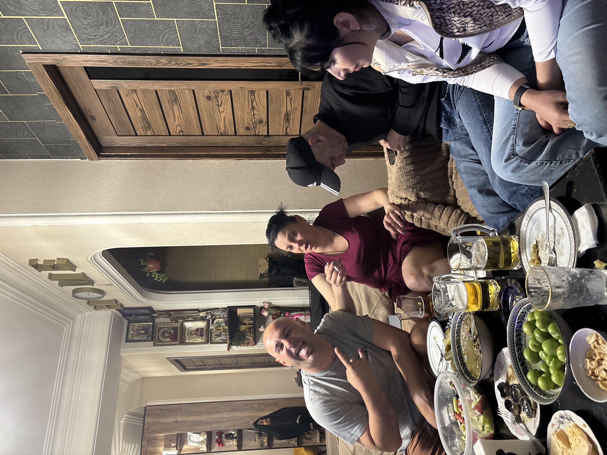 This image captures a lively indoor social gathering, likely an evening meal or celebration, with several adults congregated around a low table laden with food and drinks. The perspective is from slightly above, looking down into the scene.
At least five individuals are visible. In the bottom center, a bald man in a grey t-shirt smiles broadly at the camera, making a "rock on" hand gesture. To his right, a woman in a maroon t-shirt is seated, appearing to hold a cigarette, looking towards the man. Further up and to the right, a man wearing a black long-sleeve shirt and a black baseball cap with the Adidas logo looks downwards. Beside him, a woman in a white patterned vest and light blue jeans is seated, her hands clasped. Another person's legs in brown shorts or pants are partially visible in the bottom left.
The dark-colored table is central to the activity, covered with various dishes. These include bowls of green olives, dark olives, shelled nuts (possibly peanuts or almonds), salads in clear containers, and plates holding what appear to be dips or spreads, and possibly a dessert or cheese. Beverages include two large glass mugs filled with a light-colored liquid, likely beer, and several empty glasses and plates, indicating ongoing consumption.
The setting appears to be a cozy home environment. A plush, textured brown sofa or cushion provides seating for some of the guests. In the background, there's a wooden cabinet or door with vertical slats. The walls feature a distinctive dark grey tiled pattern with yellow or gold lines and circular motifs. The ceiling is white with decorative molding, and some three-dimensional letters, possibly spelling out a word like "HOME," are affixed to it, with "H" and "O" being most discernible. A shelf against a back wall displays numerous framed pictures, possibly religious icons, and a small potted plant. The overall mood conveyed is one of relaxed enjoyment and camaraderie.