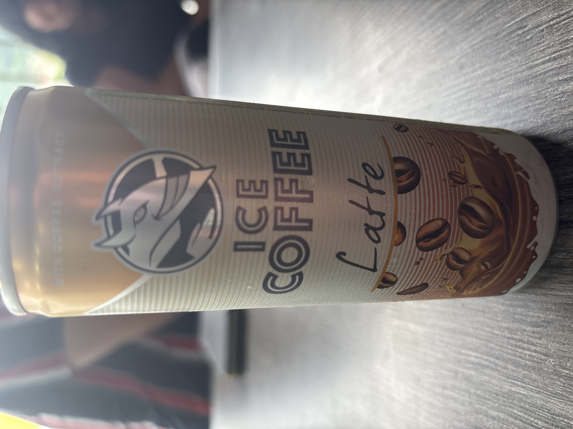A close-up, slightly angled shot showcases a can of "ICE COFFEE LATTE" with a distinctive logo of a stylized creature. The can is primarily gold and white, with thin horizontal lines creating a textured background on the white sections. The logo, featuring a gray creature with sharp features and possibly horns, is centered on the upper portion of the can. Below the logo, "ICE COFFEE" is printed in large, dark gray block letters, followed by "Latte" in a handwritten-style font. The lower part of the can displays an illustration of coffee beans splashing into a chocolate-colored liquid, with white splashes. Small water droplets are visible on the surface of the can, suggesting it is cold. The can rests on a gray, wooden surface with visible grain. In the blurred background, two individuals are partially visible. One, on the left, wears a red and black striped shirt. The other, on the right, has dark hair and wears a dark t-shirt. The overall setting appears to be indoors, possibly a cafe or restaurant, as indicated by the table surface and the blurred background figures, and the lighting suggests daylight filtering in. The specific location is the city of Karlovo, Bulgaria, though no explicit markers confirm this within the visual frame itself, this information is inferred from external context.