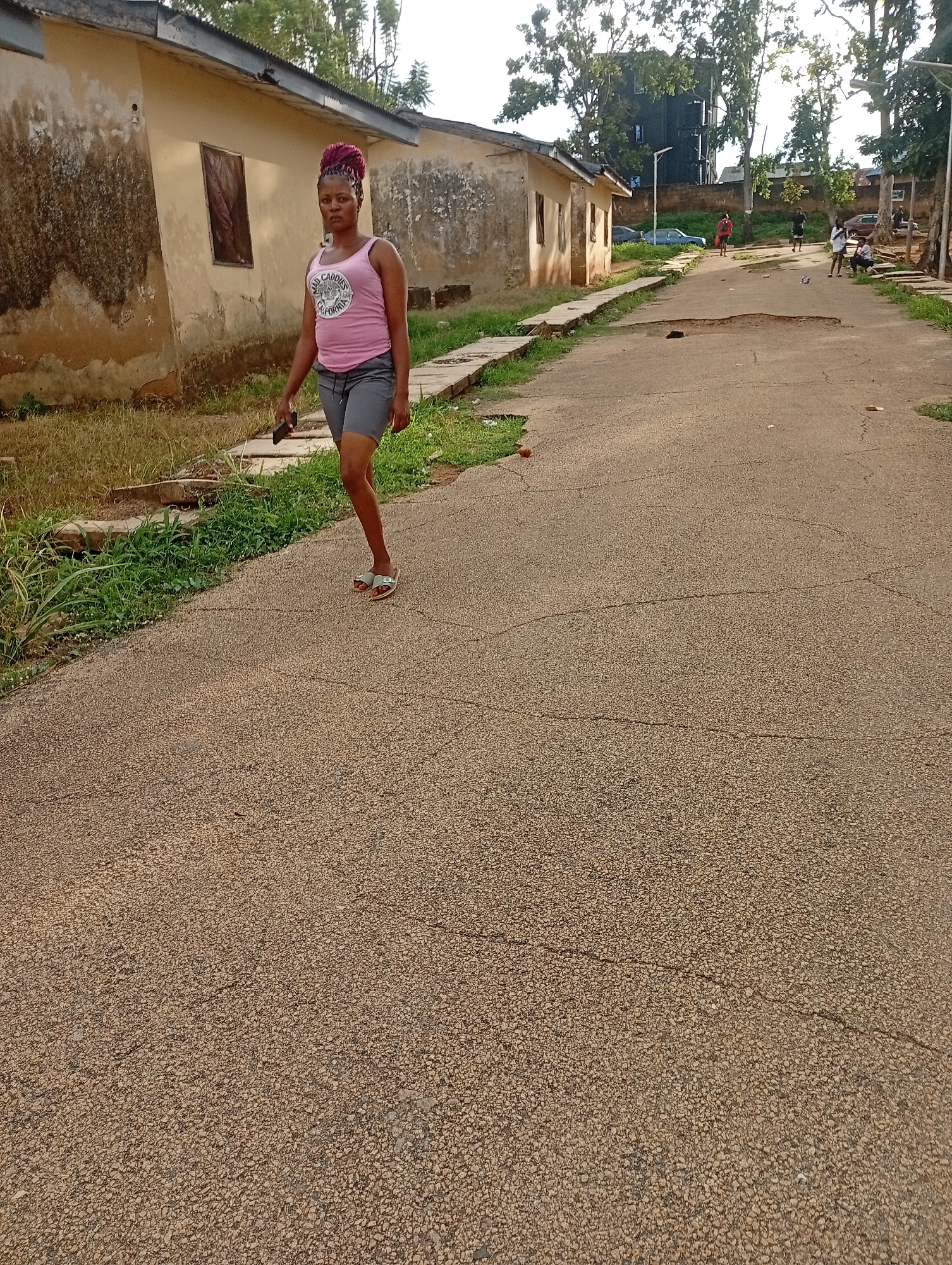 A woman with braided, purple-dyed hair stands on an asphalt road in Rafin Par, Nigeria. She wears a pink tank top with a graphic print and gray shorts, and is holding a black smartphone in her right hand. Her feet are adorned with light-colored flip-flops. She is looking directly at the camera with a neutral expression.
The scene appears to be outdoors in a semi-urban or rural setting. To the left of the woman are dilapidated, light-colored buildings with weathered walls and dark-framed windows. The buildings are lined with overgrown grass and concrete pathways. Further down the road, other people are visible. Two individuals are standing, one wearing a red shirt and the other a dark shirt. To the right, a group of three people are seated, and a soccer ball rests on the ground nearby. Cars are parked along the side of the road in the distance.
The road itself is cracked and uneven, indicating wear and tear. The presence of lush green grass and trees suggests a warm, likely tropical, climate. The lighting indicates it is daytime, possibly late afternoon or early morning, as the sun casts soft shadows. The overall atmosphere seems casual and everyday, with people engaged in various activities on the road. The graphic on the woman's tank top reads "Bad Caddies California".