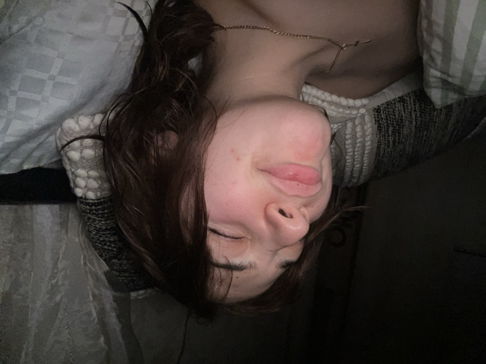 A young woman lies on her side, possibly asleep, with her eyes closed and a slight smile. She has brown hair and fair skin. A thin gold chain adorns her neck. She's surrounded by pillows and bedding. The lighting is dim, and the image is rotated, giving a slightly disorienting perspective. The overall scene appears peaceful and relaxed.