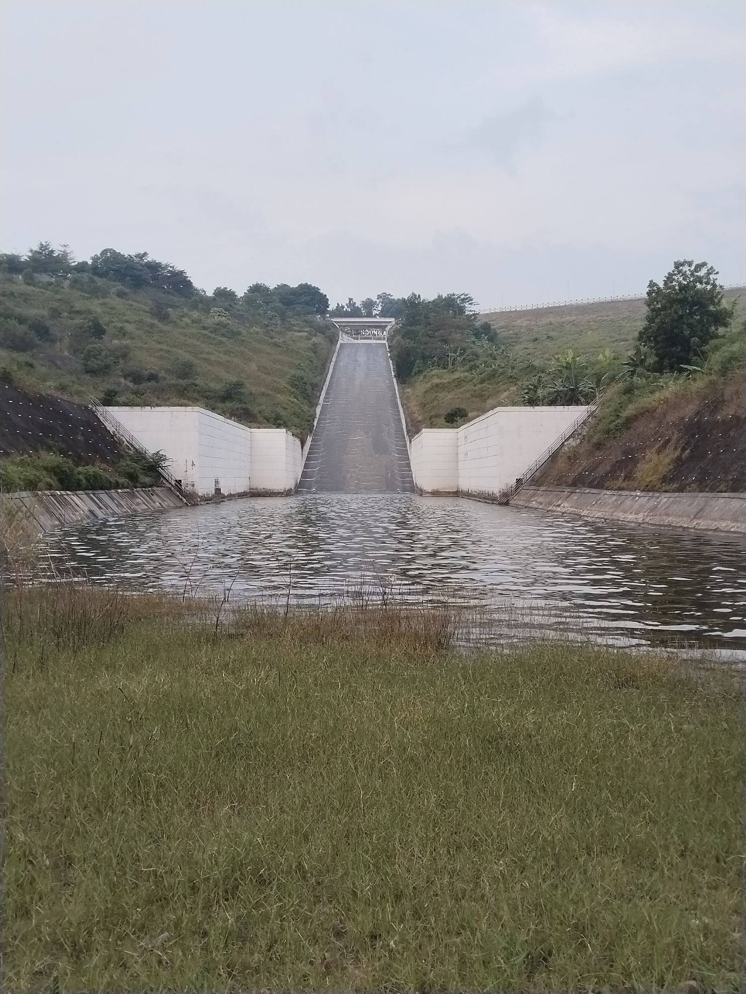 This photograph shows a water reservoir and spillway located in Ganten, Indonesia. The spillway is a long, inclined concrete structure with a bridge-like feature at its crest. The concrete structure is flanked by white, block-like walls, which transition into concrete-lined water channels. The reservoir surface is rippled, and the foreground consists of a grassy area. Vegetation covers the surrounding slopes. The sky is overcast.