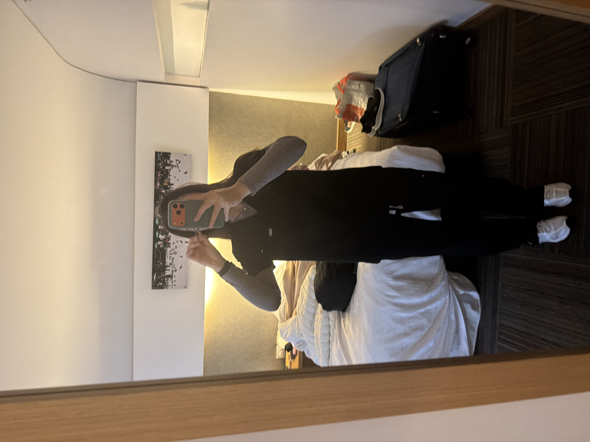 A person stands in a hotel room, reflected in a large mirror. They are wearing dark scrubs, a grey long-sleeved shirt underneath, and white sneakers. The person is holding a smartphone in their right hand and appears to be adjusting something near their ear with their left hand, possibly an earbud. The room has a bed with white linens, a bedside table, and a dark suitcase with a canvas bag on top. The walls are cream-colored, and a piece of art depicting a flock of birds in a winter scene hangs above the bed. The flooring is a patterned carpet in shades of brown and grey. The overall impression is of a tidy but functional hotel room. The image does not provide any specific visual cues to pinpoint the location within Bogota, Colombia.