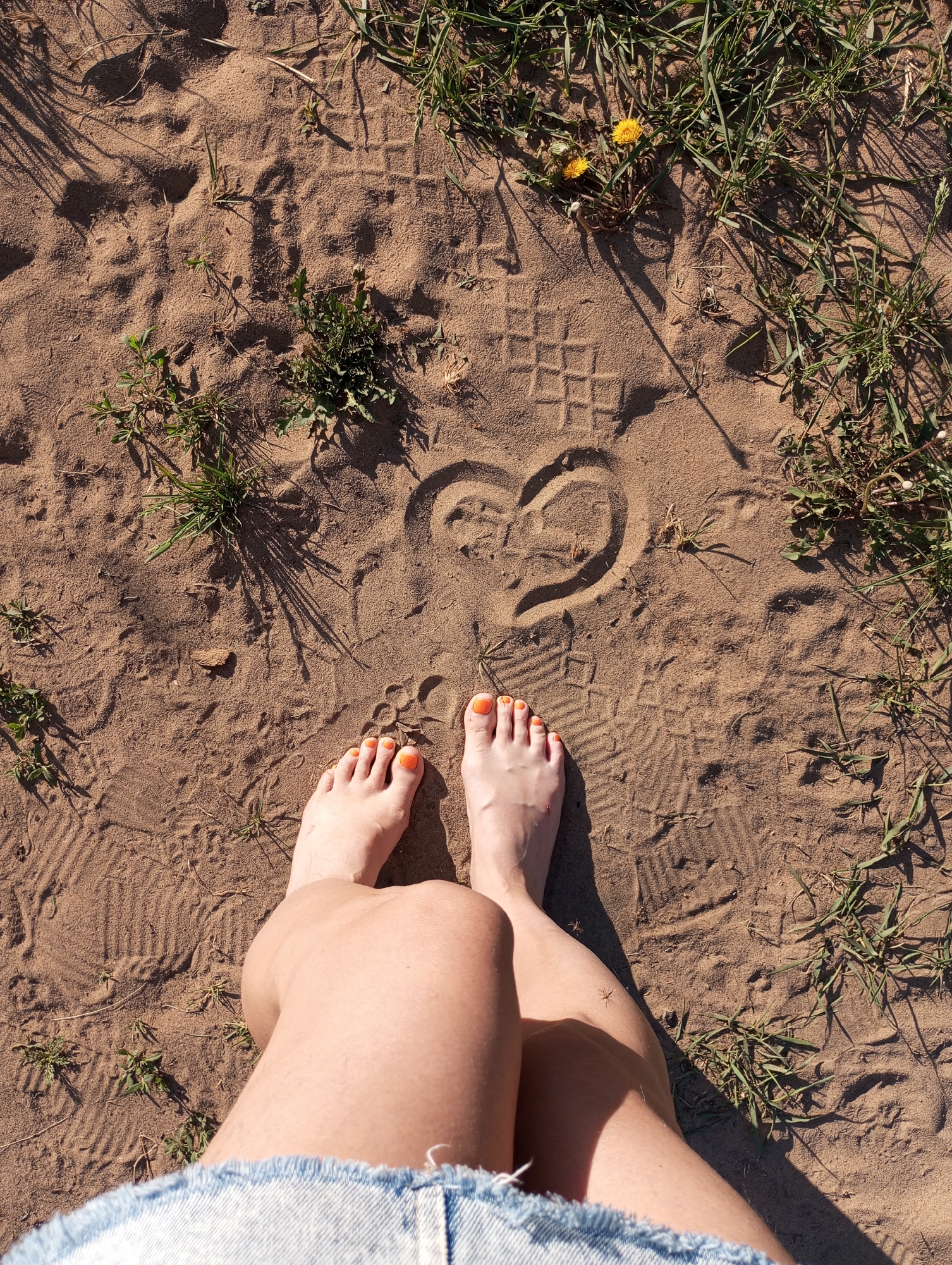 A vertical, full-body shot depicts a person standing on a sandy area with patches of grass. The individual is wearing denim shorts and has bare feet with orange nail polish on their toes. They are standing in front of a heart shape drawn in the sand, surrounded by footprints. Patches of grass and small yellow flowers are visible. The photo seems to be taken during the day with natural lighting. The location cannot be determined to be Буренка, Russia, based on the image alone.