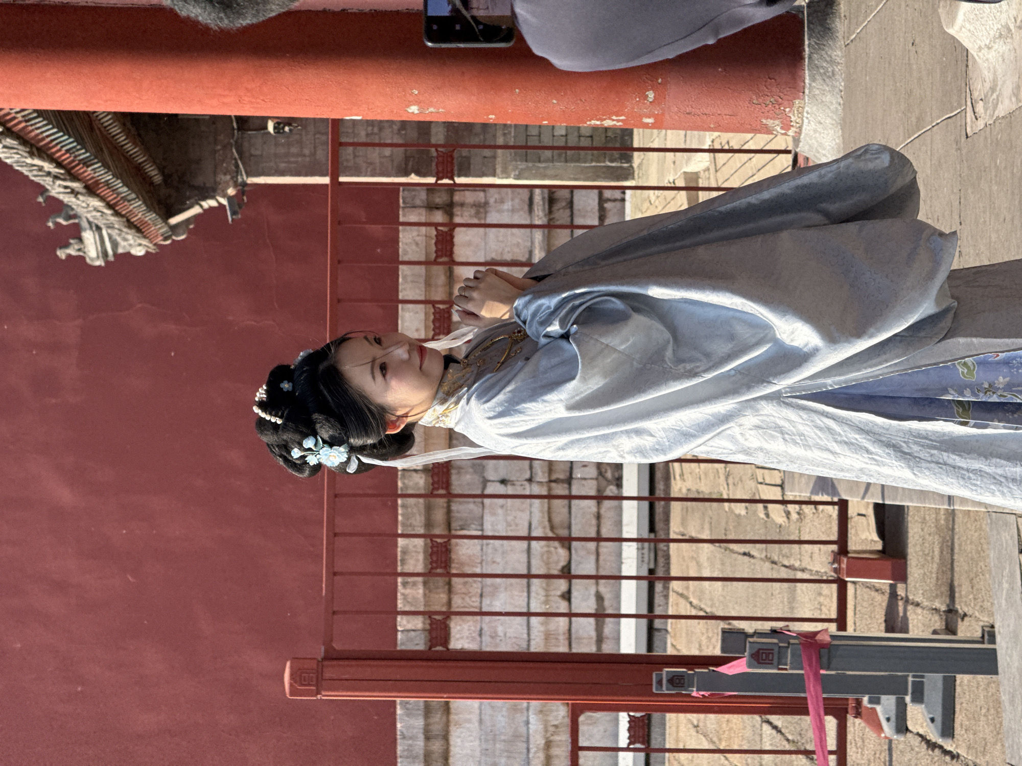 The image shows a woman dressed in traditional Chinese Hanfu clothing standing in front of a red wall with a railing. The location appears to be in the Dongcheng District of China, based on the architectural style. The woman has her hair styled with traditional ornaments. A portion of another person is visible in the upper left corner, and a smartphone is also visible. The railing is made of red-painted metal with a horizontal bar design. The wall is a dark red color, likely part of a traditional building. There is a modern-looking grey fixture with a red ribbon tied around it near the bottom right corner of the image. The ground is paved with stone.