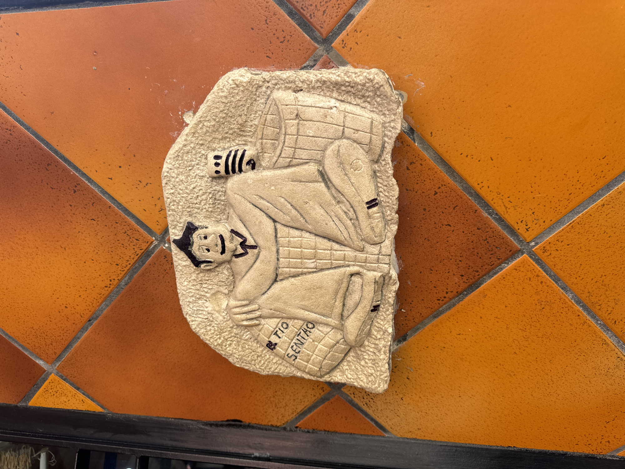 This is a close-up, eye-level, medium shot of a bas-relief carving depicting a seated man in traditional clothing, likely part of a decorative facade. The carving is light-colored and textured, contrasting with the glossy, orange, diamond-patterned tiles of the wall it's mounted on. The man has a smiling expression, a dark cap, and is holding a bag with text that reads "TIO SENTAO" on his left. To his right is a woven basket. The wall tiles are arranged in a diagonal grid with dark grout lines. A dark, vertical frame runs along the left edge of the image. The overall scene appears to be outdoors, given the texture and color of the tiles, which are common in Spanish architecture. Without further context or identifiable landmarks, the specific location in Murcia, Spain, cannot be determined.