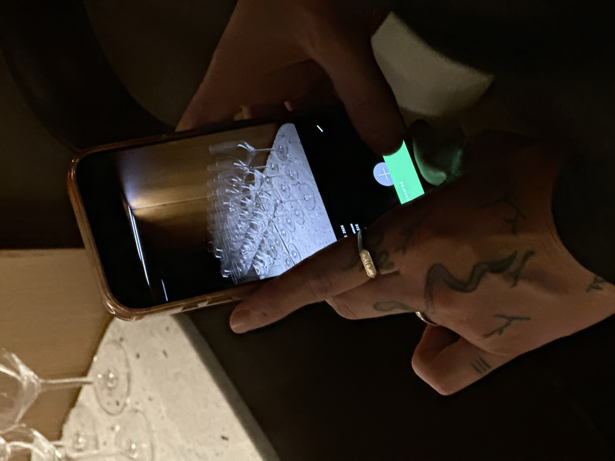 A close-up shot shows a person holding a smartphone. The phone's screen displays an image of numerous wine glasses arranged on a surface. The person's hand, adorned with tattoos and a ring, is operating the phone. Some wine glasses are also visible in the foreground outside of the screen. The setting seems dim, possibly indoors. The location is inferred to be Yerevan, Armenia.