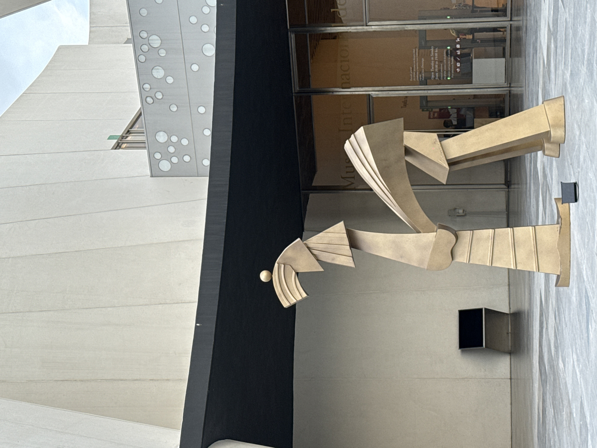 A bronze sculpture stands outside a building in San Bernardino Tlaxcalancingo, Mexico. The sculpture is composed of geometric shapes. The building is modern with white panels and a black facade. The building's glass doors show the words "Museo Internacional del Barroco".