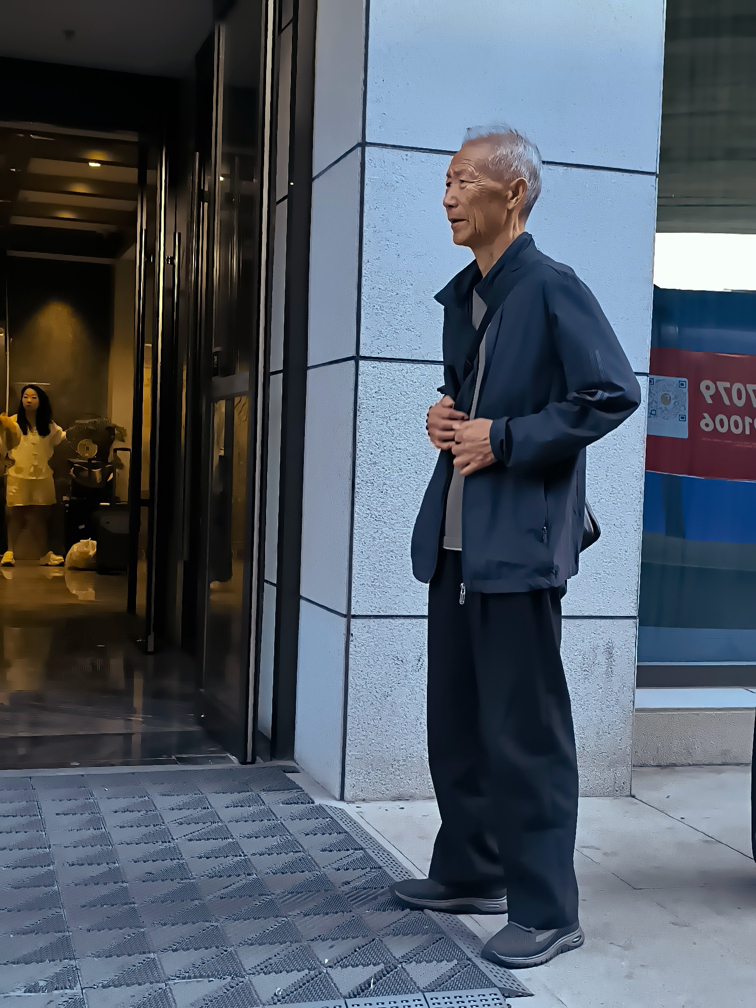 An elderly Asian man with gray hair stands on a patterned walkway in front of a modern building. He is wearing a dark jacket over a light-colored shirt and dark pants. His hands are clasped in front of him. To his left, through an open doorway, a young Asian woman in a yellow outfit is visible inside a dimly lit building. The building exterior features a textured facade. The scene appears to be outdoors in an urban environment. The presence of a patterned walkway suggests a public or commercial area, possibly designed for accessibility. The background includes a glimpse of a blue and red sign on another structure, partially obscured. The overall lighting suggests it is daytime, though the interior of the building is dark. The man's posture and gaze indicate he is looking to his right, towards the building's exterior.