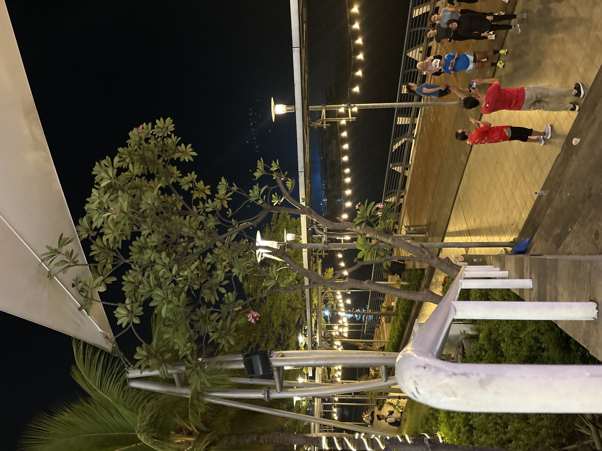 The image depicts an outdoor night scene at Ancol Beach City, North Jakarta, Indonesia. A group of people are gathered on a walkway near a body of water, illuminated by string lights and streetlights. Some individuals appear to be exercising or preparing for an activity, with a few holding phones, possibly recording or taking pictures. The environment features palm trees, lush greenery, and a modern architectural structure with a large canopy overhead. The overall atmosphere suggests a recreational or public gathering space.