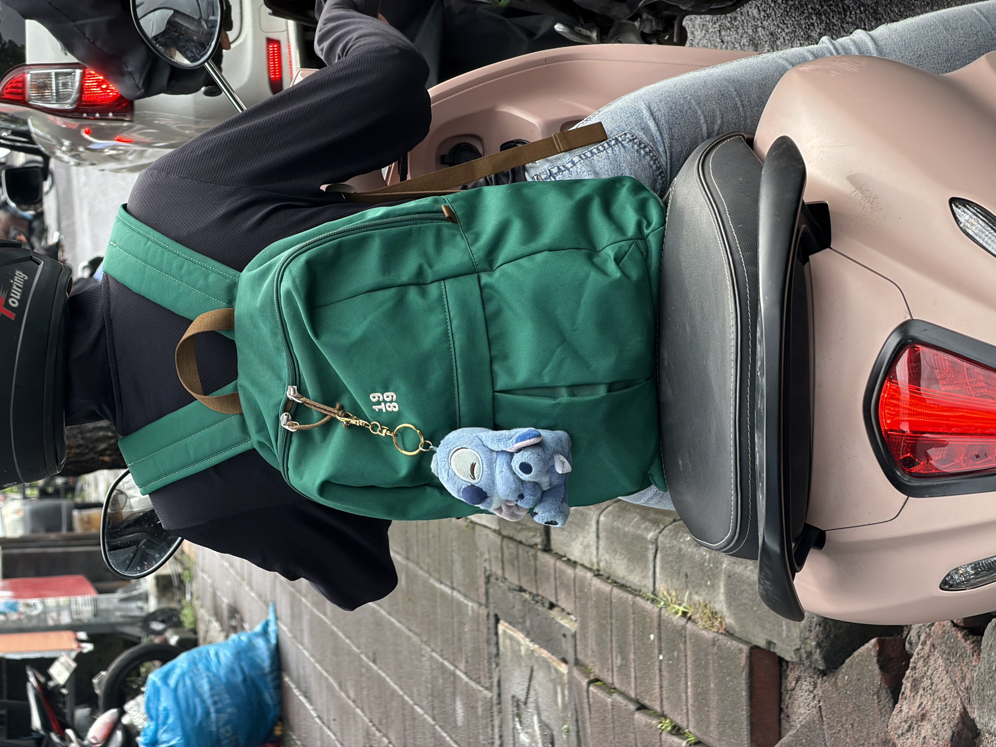 The image captures a person from a slightly elevated, side-rear perspective, seated on a light pink scooter on what appears to be a street or alleyway in North Kuta, Indonesia. The person, viewed from behind, wears a black long-sleeved top and light blue jeans. A prominent dark green backpack, featuring brown straps and a canvas-like texture, rests on their back.
Attached to the backpack is a blue plush keychain resembling the Disney character Stitch, along with a small, golden metal tag embossed with the numbers "19 89". Part of a black helmet is visible on the left, with the word "Touring" partially discernible on it.
The scooter has a black seat and a distinctive light pink or beige body. Its red taillight and a chrome rearview mirror are visible. The background shows glimpses of other vehicles, likely motorcycles or scooters, and a grey brick wall or curb running along the side of the street. A blue plastic bag or object lies on the ground near the wall. The scene is brightly lit, suggesting daytime. The overall image is rotated 90 degrees clockwise.