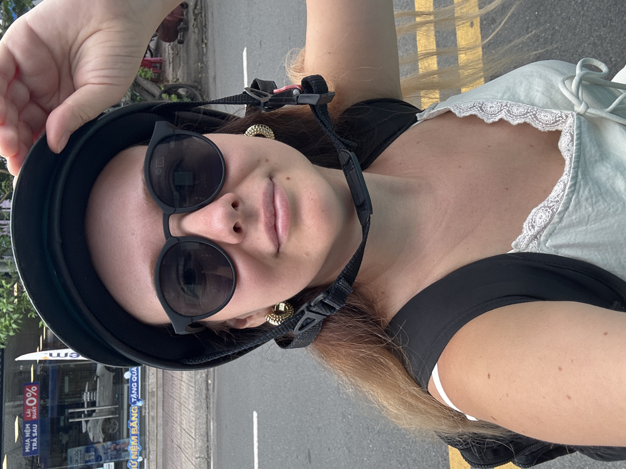 A woman, wearing a black helmet, sunglasses, and a white top with lace trim, takes a selfie outdoors in an urban environment. She is holding the brim of her helmet with her right hand. The background shows a street with yellow lines, a sidewalk, and a building with signage that includes "EM" and "0%". Her left arm is visible, as is her blonde hair. The overall scene suggests a casual outing in a city.