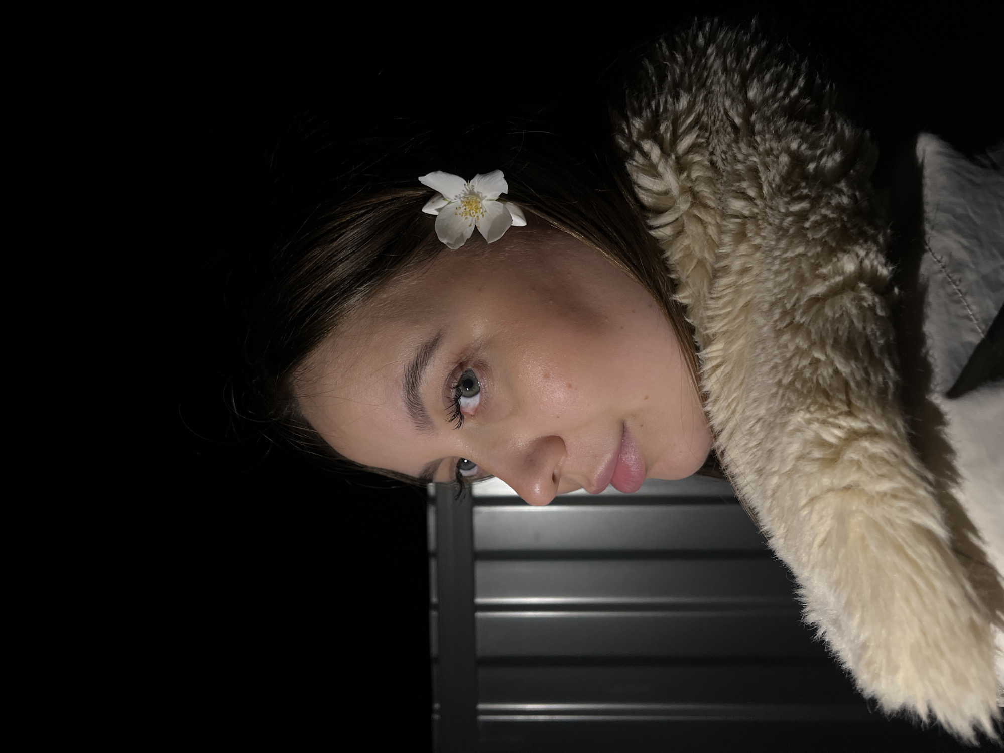 The image presents a close-up, tilted portrait of a young female, likely an adult or older teen, captured at night or in a dimly lit environment. Her head is tilted significantly to the right, with her face looking upwards and slightly towards the viewer. She has brown hair, fair skin, and light-colored eyes. A small, white five-petaled flower with a yellow center is tucked into her hair above her right eye.
Her expression is serene and gentle, with a soft, subtle smile on her lips. She wears a garment with a prominent, fluffy, cream-colored faux fur collar or hood lining, visible along the right side of the frame and partially obscuring the top of her head. The background is predominantly dark, almost entirely black, indicating nighttime or a deeply shadowed setting. In the lower right portion of the image, a dark, horizontally ribbed or slatted structure is visible, suggesting a wall, fence, or set of blinds.
The lighting appears to be focused on her face, highlighting her features against the dark surroundings. No specific activity is taking place beyond posing for the portrait. The overall mood is calm and intimate. There is no visible text in the image. The location context is Novopokrovskaya, Russia.