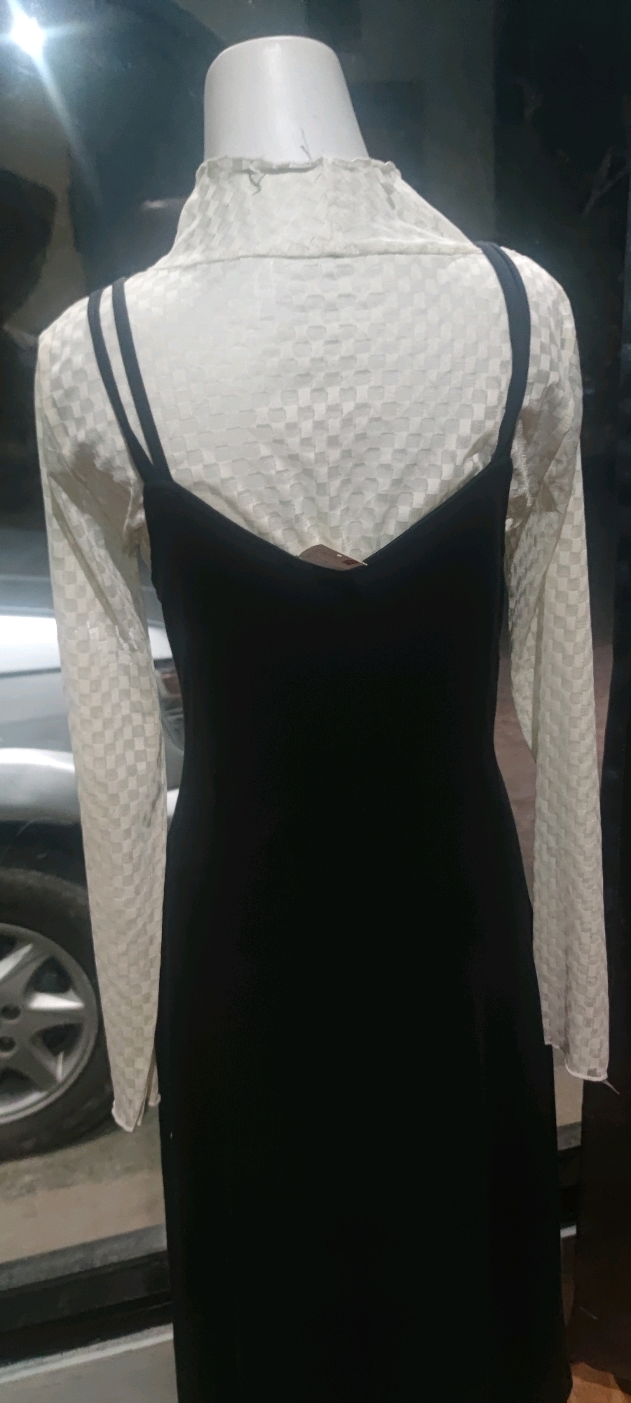 The image shows a mannequin wearing a layered outfit in what appears to be a retail setting. The mannequin is dressed in a white, high-necked, long-sleeved blouse with a subtle checkered pattern. Over the blouse, a black slip dress with thin straps is worn. The background is dark and somewhat blurry, suggesting it might be a shop interior. On the left side of the image, a portion of a car tire and rim is visible, indicating the presence of a vehicle nearby, possibly outside the shop or in a parking area. The overall lighting is dim. There is no visible indication of the location being Gambarsari, Indonesia.