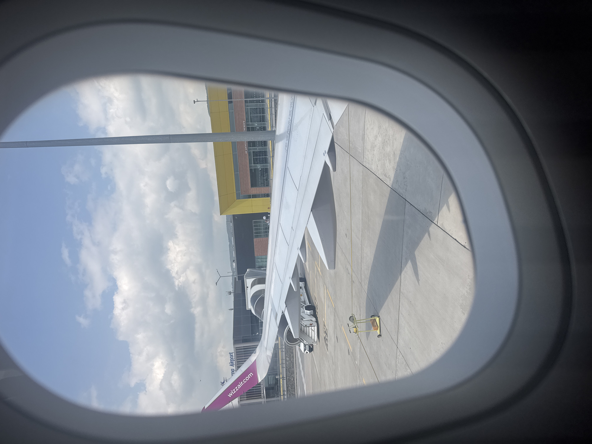 The photograph, taken from inside an airplane window, captures a scene at an airport during daytime. The view is dominated by the white wing of a Wizz Air aircraft, which extends from the bottom left towards the center and right. The airline's purple branding and "wizzair.com" logo are visible on the underside of the wing near the fuselage.
Beyond the wing, an airport terminal building is visible. It features a distinct yellow upper section with large glass windows, and a darker lower facade. Attached to the building, a jet bridge or air stairs are positioned near the aircraft's door, suggesting either boarding, disembarking, or preparation for departure/arrival. Various ground service equipment, including a yellow baggage cart, is seen on the grey tarmac beside the aircraft.
The sky is bright blue with scattered white, fluffy clouds, indicating clear weather. The sun is high, casting distinct shadows of the aircraft wing and other structures on the tarmac. A tall, slender light pole is also visible against the sky.
Visible text on the terminal building includes "НЕ ШЕНГЕН" (Non-Schengen) on a brown-toned sign, and "BURGAS AIRPORT" on a white sign below the yellow section. Another larger sign, partially visible, reads "Wizz Air" and "Airport". The location context confirms this is likely Burgas Airport in Aksakovo, Bulgaria. The overall impression is that of an active airport environment under sunny conditions.