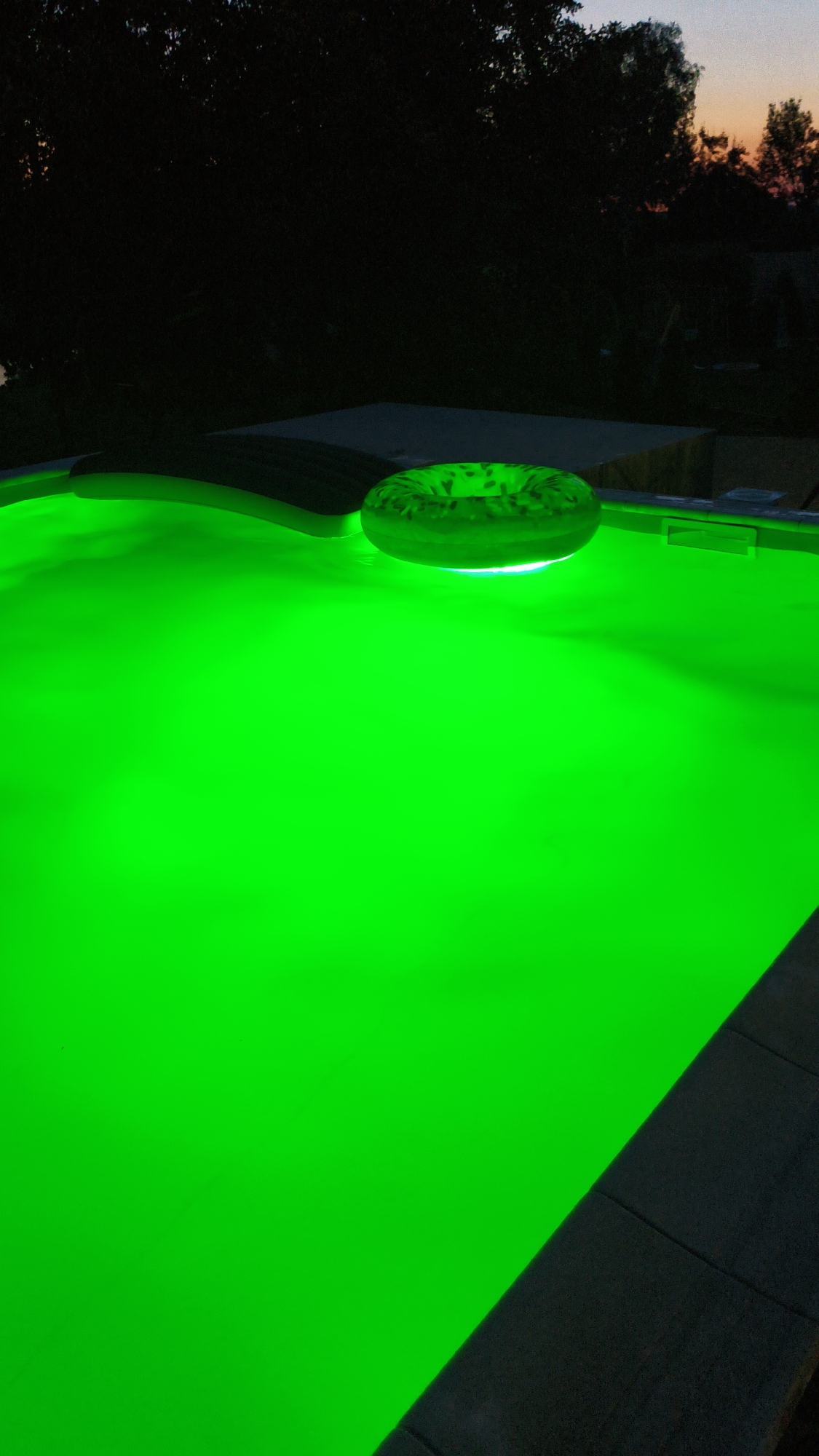 An illuminated swimming pool in Bezliudivka, Ukraine, glows a vibrant green at dusk. A green inflatable ring with a patterned surface floats on the luminous water. Dark trees and a dusky sky are visible in the background. The pool's edge, made of grey concrete pavers, is partially visible in the foreground and along the right side.