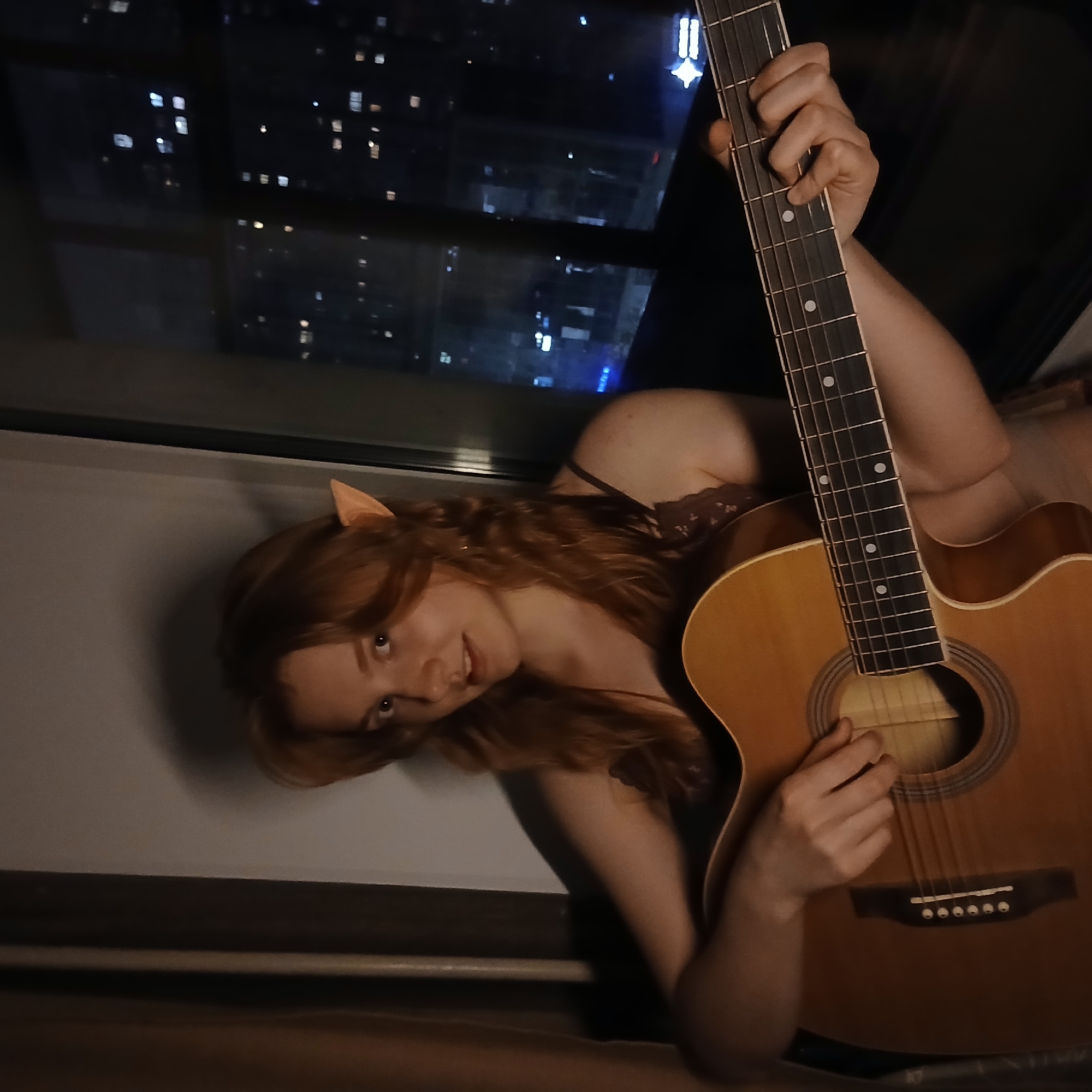 A young woman with long, reddish-brown hair is depicted indoors at night, reclining by a window overlooking a city. She wears small, light-colored ear-like prosthetics, resembling elf ears, and a dark, possibly lace-trimmed, top. Her expression is calm with a slight smile as she looks towards the viewer.
She is holding a light-colored acoustic guitar, with her left hand positioned on the fretboard and her right hand near the soundhole, suggesting she is playing or posing with the instrument. The room is dimly lit, contrasting with the vibrant cityscape visible through the window. Numerous illuminated building lights, appearing as bright squares and points, punctuate the dark night sky outside, indicating an urban environment like Yekaterinburg, Russia. The city lights reflect faintly on the windowpane. The image is rotated, giving the impression that the woman is lying on her side or back with the window above her. There is no visible text in the image.