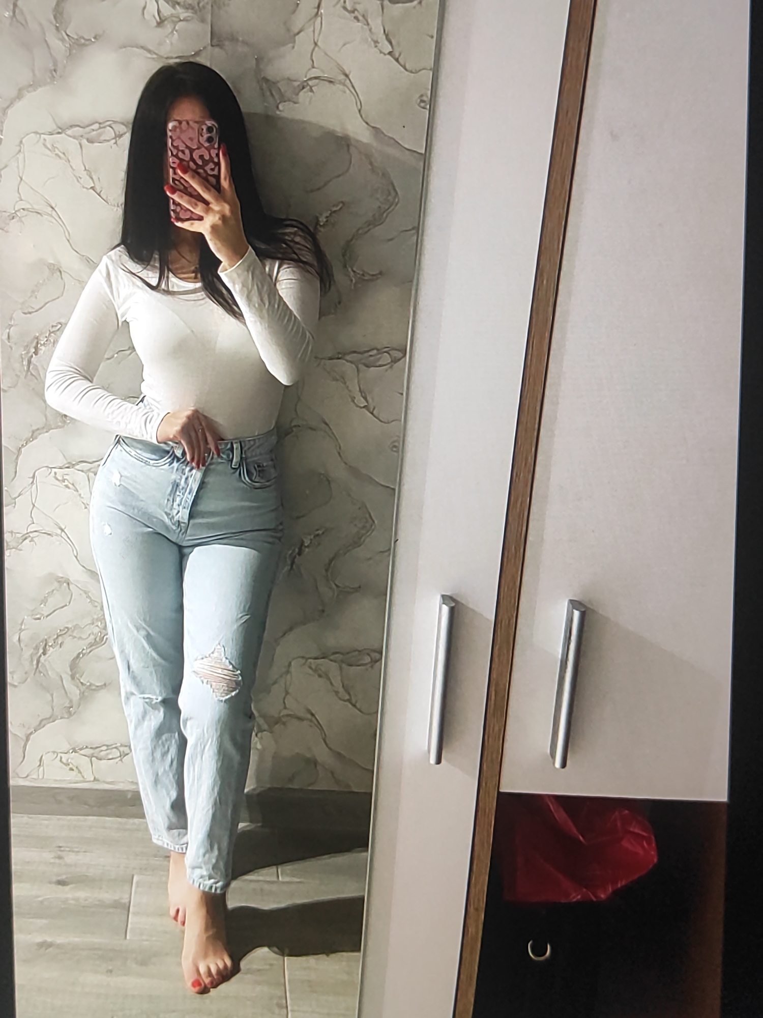 In this media file, a young woman is captured in a mirror selfie. She is standing in what appears to be a bedroom or dressing area, with a marble-patterned wall behind her and a large wardrobe to her right. The woman is wearing a white long-sleeved shirt and light-wash ripped jeans. She is holding a smartphone in her right hand, which is partially obscuring her face. Her left hand is resting on her hip, adjusting her jeans. Her toenails are painted red.
The scene is well-lit, suggesting it is daytime or that the room has good artificial lighting. There are no clear indicators of the weather. The location cues are minimal, but the presence of a wardrobe and the casual attire of the woman suggest a domestic setting. The overall emotion conveyed is one of self-assurance and personal style. There is no visible text in the media file.