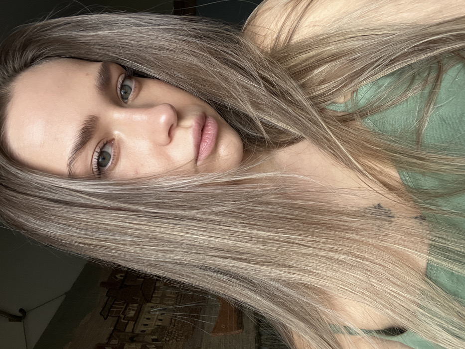 The image displays a close-up portrait of a single female subject, likely taken indoors within a residential-like setting in Saint Petersburg, Russia. The subject is a young adult with long, straight, light brown hair featuring blonde highlights, which partially covers her shoulders and upper chest. Her light-colored eyes are directed towards the camera, and her expression is neutral. She is wearing a light green tank top. A small, dark tattoo, possibly a stylized star or abstract shape, is visible on her upper left chest, partially obscured by her hair. The background is out of focus, revealing a tapestry or wall art on the left side, depicting an old European coastal town scene with buildings and boats in muted tones. No specific landmarks are visible to pinpoint the precise location within Saint Petersburg beyond an indoor domestic environment.
