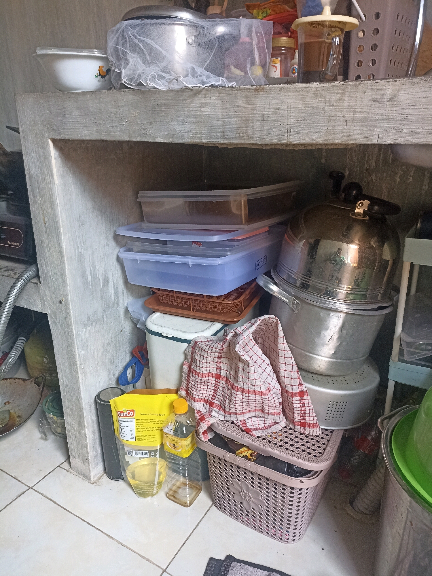 The image shows a cluttered kitchen shelf. On the top shelf, there is a white bowl with a rooster design, a large pot covered with a white net, and a glass containing a dark liquid, possibly coffee or tea, with a yellow lid. To the right, a gray wire basket is visible.
The middle shelf contains several clear plastic containers stacked on top of each other, with a brown woven basket underneath the top two. Below these, there is a white and green rectangular container.
On the bottom shelf, a large aluminum pot is stacked with what appears to be a steamer insert on top. A red and white checkered cloth is draped over a beige plastic basket, which contains various items, including a black plastic bag. To the left, a yellow packet of "SunCo" cooking oil is standing upright, next to a partially filled plastic bottle of cooking oil. A roll of gray tape is also on this shelf.
The kitchen appears to be in a residential setting, possibly in Mojopanggung, Indonesia, given the context. The overall impression is one of a functional but untidy kitchen space.
