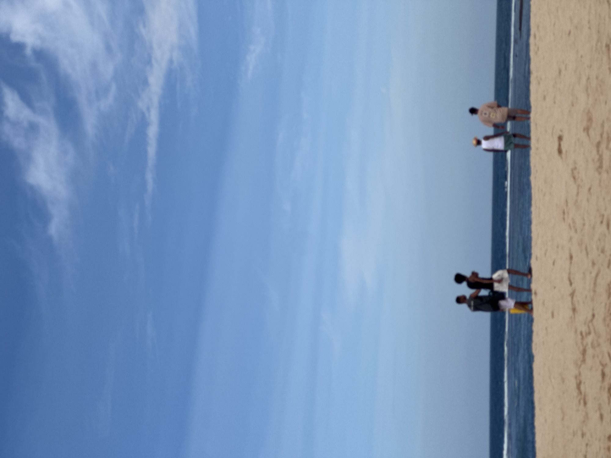 The image depicts a beach scene in Kuta Selatan, Indonesia, under a clear blue sky with wispy clouds. The foreground is a sandy beach, leading to the ocean with gentle waves. In the mid-ground, four individuals are visible standing on the beach, facing the ocean. Two men on the left are dressed in dark shirts and light shorts, with one holding a yellow object. Two individuals on the right are seen from behind, one wearing a white tank top and green shorts, and the other a patterned light-colored shirt and dark shorts. The overall atmosphere is serene and suggestive of a peaceful day at the coast.