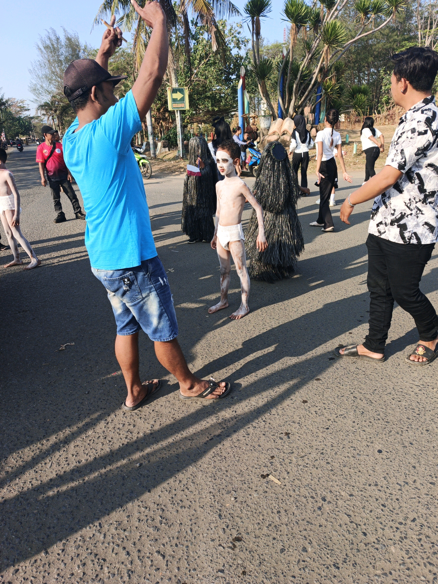 In Kelurahan Cilacap, Kecamatan Cilacap, Indonesia, a street scene unfolds under a bright sky, casting long shadows across the asphalt. The foreground is dominated by a person in a bright blue t-shirt and denim shorts, their arms raised and one leg extended, appearing to be directing or performing. To their left, a young child, their skin painted white and wearing white briefs, stands with their arms at their sides. Behind this child are two figures in tall, dark, shaggy costumes, one of whom is holding a small Indonesian flag. Further back on the left, a person in a red shirt and black pants walks away from the camera. To the right, a person in a black and white patterned shirt and black pants walks towards the foreground. In the middle ground, several other individuals are visible, some in casual attire and others in what appear to be costumes. Palm trees and other vegetation line the street, suggesting a tropical or subtropical environment. The overall atmosphere appears to be a public gathering or event, possibly a parade or festival.