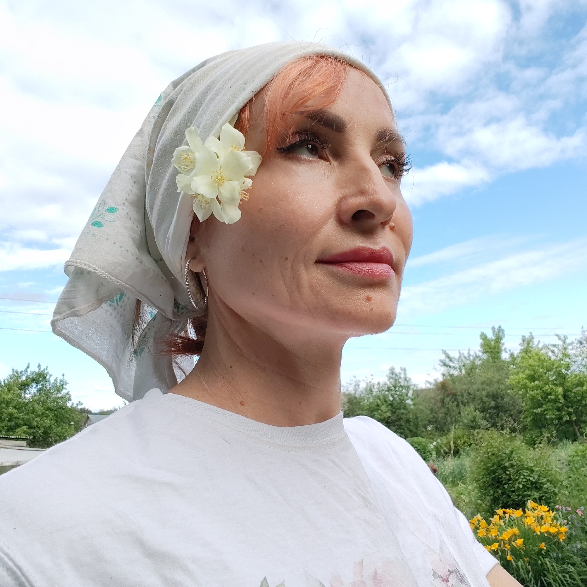 A woman with short, peachy-colored hair, wearing a white bandana and a white t-shirt, poses for a photo outdoors. A white flower is tucked into her bandana. She is looking up and to the right, with a slight smile. The background consists of green trees and a blue sky with white clouds. The scene appears to be in a rural or suburban area, possibly a garden or backyard. There are no identifying landmarks or clues to suggest the location is specifically in the city of Chaikovsky, Russia.