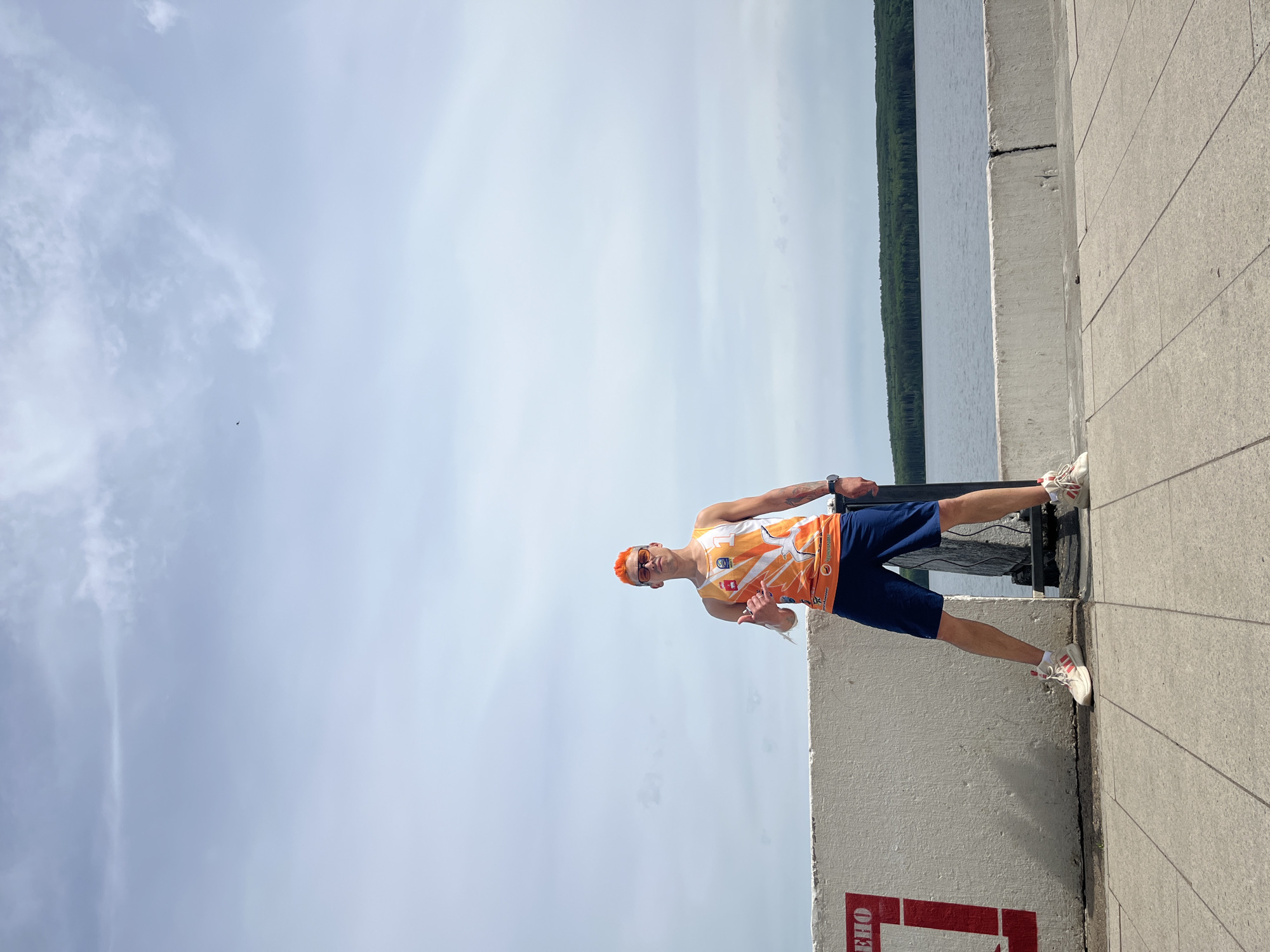 A man stands on a concrete ledge in Chaikovsky, Russia. He wears an orange tank top, blue shorts, white sneakers, an orange headband, and sunglasses. He holds something white in his hand and makes a hang loose hand gesture. Behind him are a body of water and trees. The sky is cloudy.