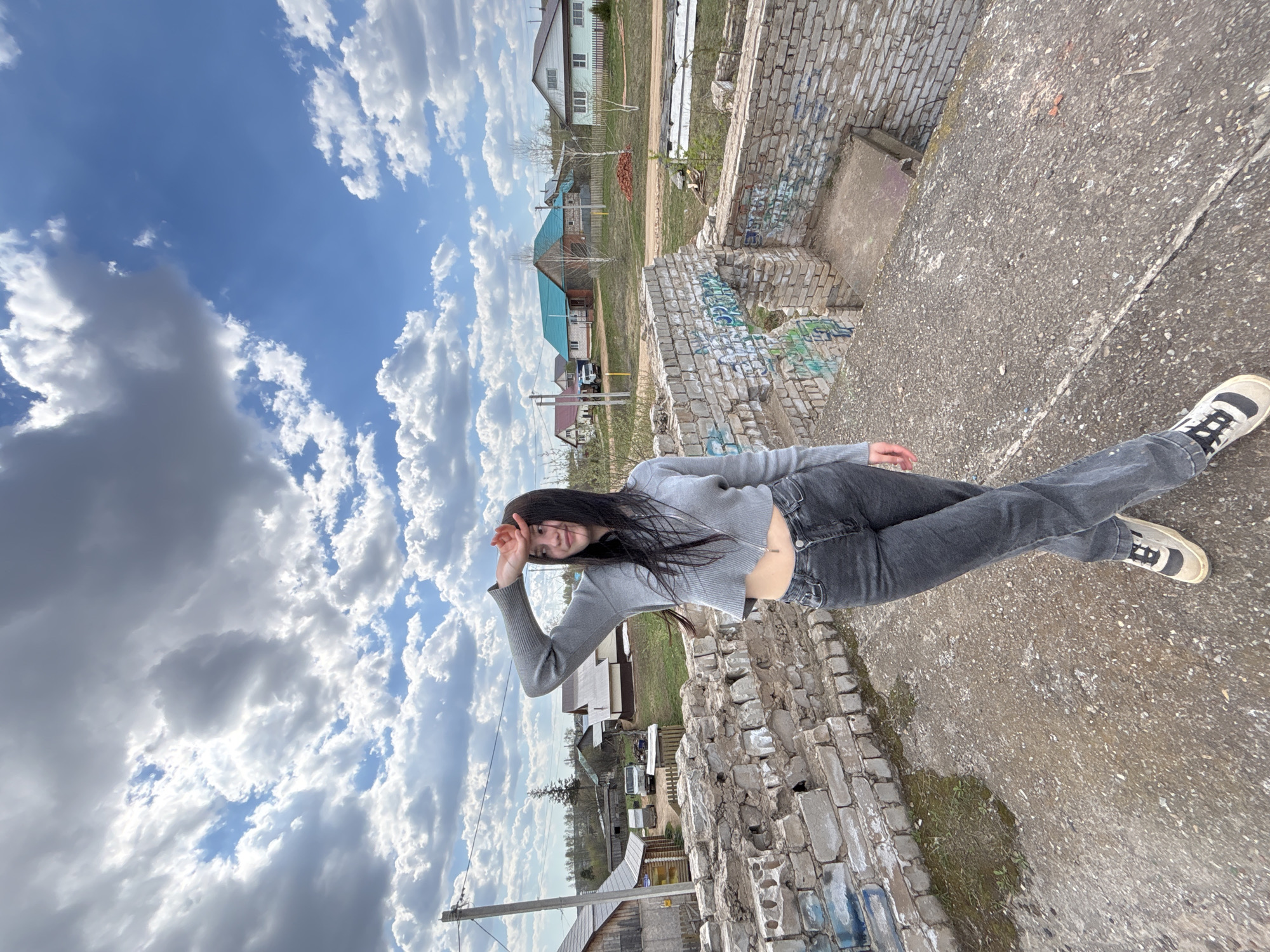 The image shows a woman standing on a concrete and brick structure in what appears to be a rural or semi-rural area. The location is identified as the city of Фоки, Russia. The woman is wearing a gray sweater, dark jeans, and sneakers with dark accents. She is holding her hand up to shield her eyes, possibly from the sun. The background includes a blue sky with white clouds, as well as houses and vegetation. There are signs of dereliction in the form of broken brick walls with graffiti.