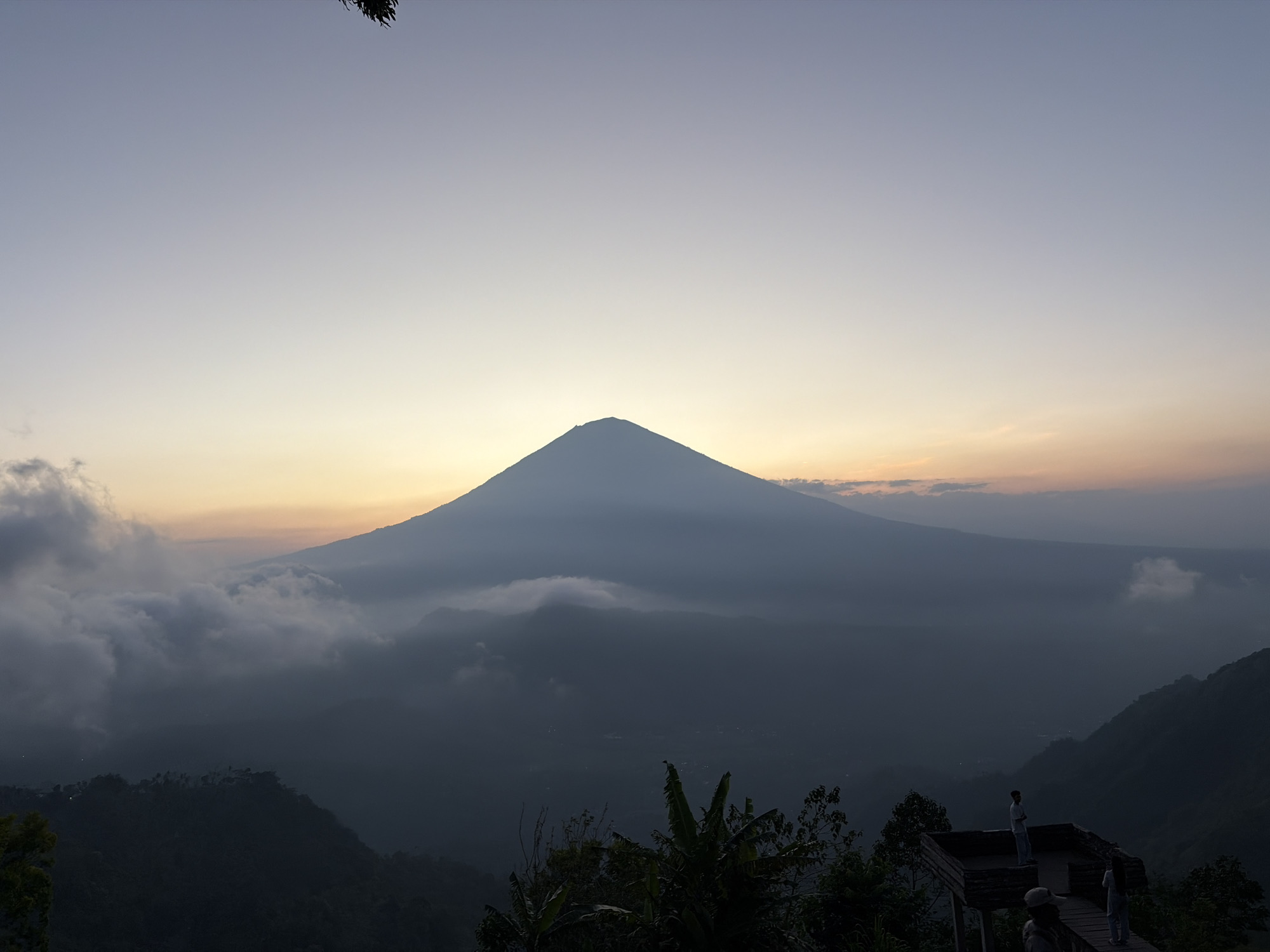 In the city of Abang, Indonesia, a majestic volcano dominates the horizon, shrouded in a soft, ethereal mist. The sky above is a gradient of pale blue to soft yellow, indicating either dawn or dusk. The peak of the volcano is sharply defined against the luminous sky, its slopes veiled in a hazy, atmospheric glow. Low-lying clouds drift around the base of the volcano, creating a dramatic, layered effect with the surrounding hills.
In the foreground, a wooden observation deck is perched on a steep incline. Two figures, appearing as silhouettes against the backdrop, stand on the deck, looking out at the panoramic view. One figure stands with hands on hips, while the other is turned away from the camera. Below them, a pathway leads to another platform where a third person, mostly obscured by shadow, can be seen. Lush green vegetation, including banana trees, frames the scene in the lower half of the image, adding a touch of tropical vibrancy to the otherwise muted landscape. The overall impression is one of serene natural beauty and a moment of quiet contemplation.