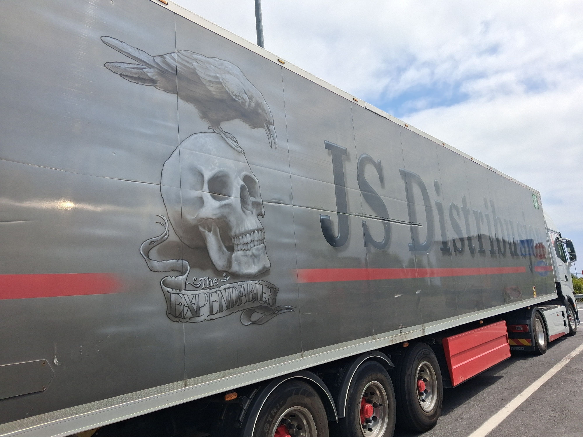 The image shows the side of a silver semi-trailer truck with a mural painted on it. The mural features a crow perched on a human skull, with a banner below the skull that reads "The Expendables". The truck has the logo "JS Distribution" painted in large, dark grey letters on its side. There is a thin red stripe running horizontally across the trailer below the mural. The truck's wheels are visible, as is part of the white cab. The truck is parked on a paved surface, possibly a road or parking lot, with a white line visible on the right side of the image. The sky is cloudy and blue. The location is identified as Bastia, Italy.