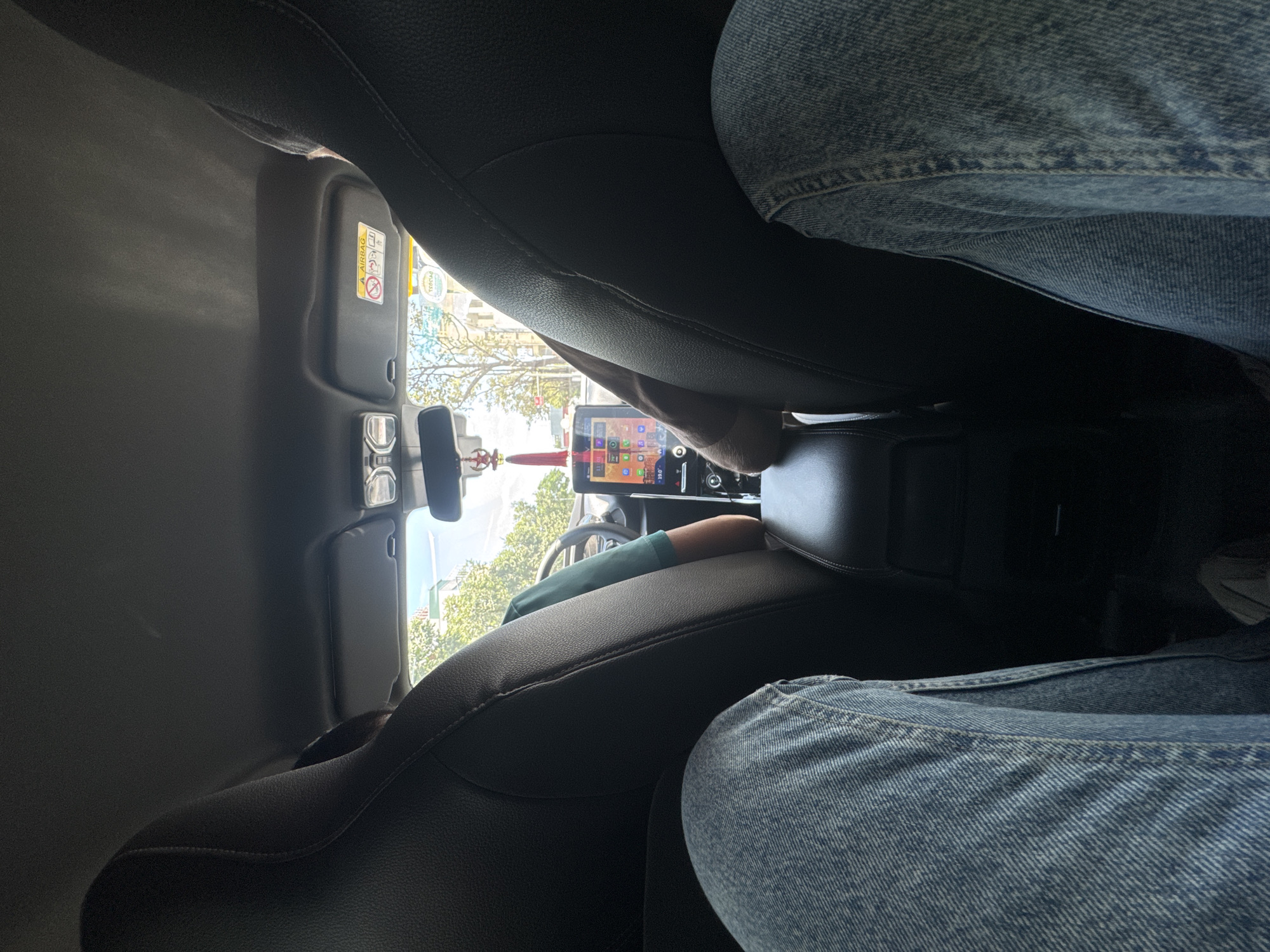 The image shows an interior view of a car, seemingly taken from the back seat. The back seats are black leather. Two pairs of legs wearing blue jeans are visible. In the front of the car, the dashboard and windshield are visible. The rearview mirror is visible. A screen is in the center console. Outside the windshield, trees and buildings are visible, suggesting an urban environment. It is difficult to pinpoint the exact location in Đà Nẵng, Vietnam.