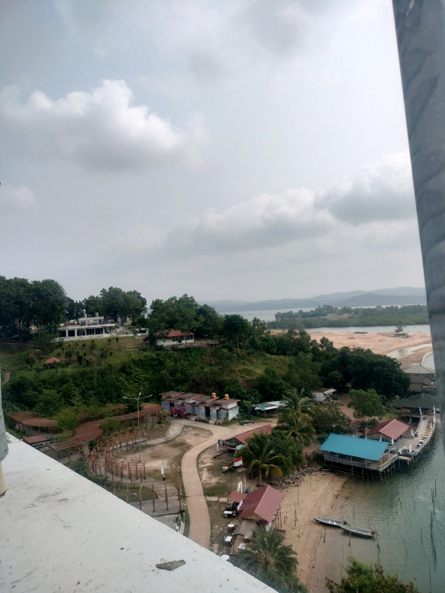 This aerial, high-angle, full shot captures a waterfront scene with buildings, vegetation, and a cloudy sky over the city of Batam, Indonesia. In the foreground, a concrete structure is visible. The middle ground features a green, hilly landscape dotted with trees and various buildings, including a white building with a sign that reads "HOTEL LANJUT". Several wooden structures with red and blue roofs are situated along the coastline, some appearing to be piers or overwater bungalows, with a small boat moored on the sandy shore. A path winds through the area, and there are outdoor seating areas with tables. In the background, a body of water stretches towards a distant, hazy shoreline with rolling hills. The sky is overcast with scattered clouds, suggesting a potentially humid day. No people are clearly identifiable in the frame.