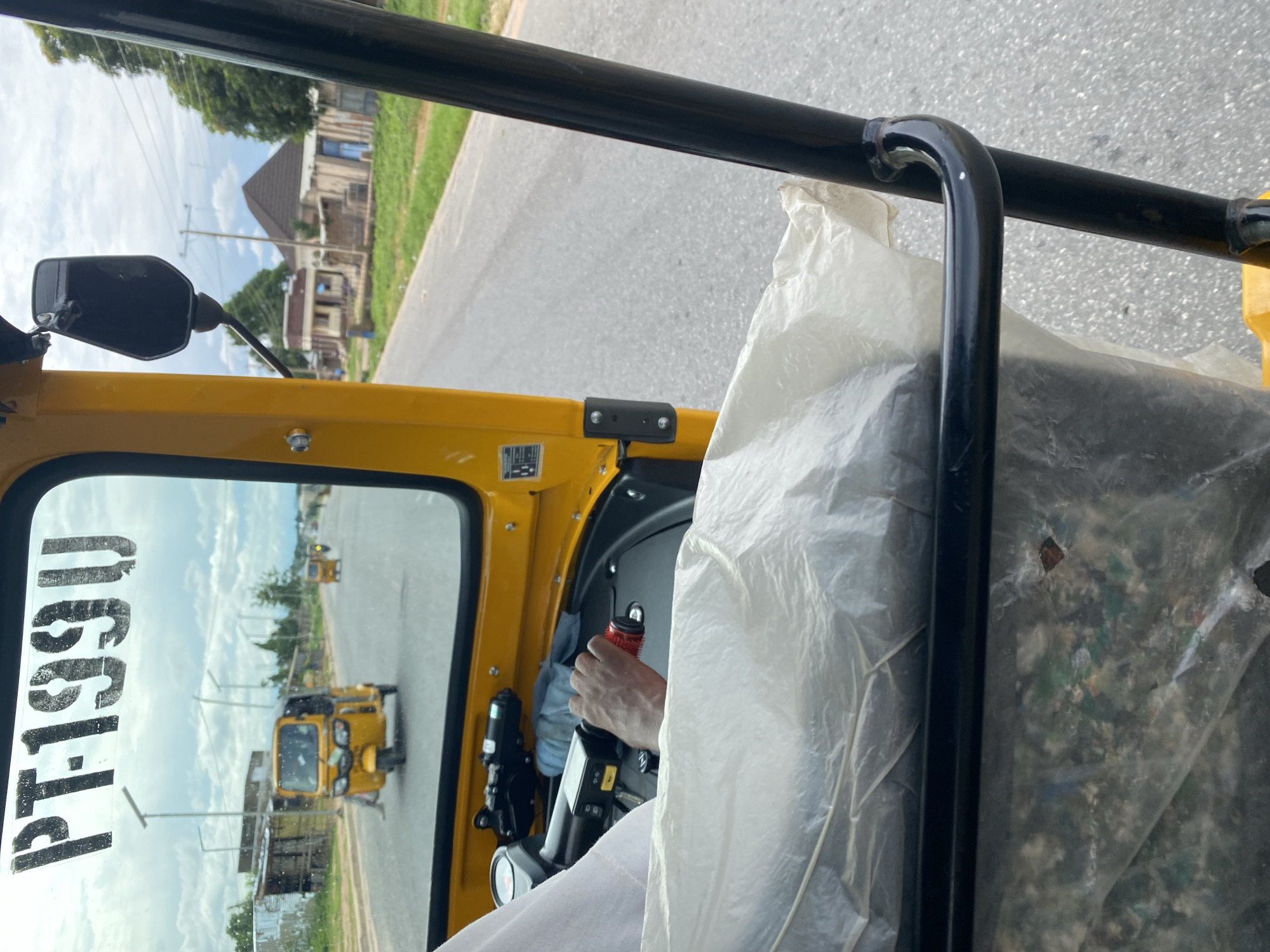 This media file captures a first-person perspective from inside a yellow auto-rickshaw, likely in Jalingo, Nigeria. The scene depicts the driver's hand, gripping a red and black lever, presumably the gear shift, indicating the vehicle is in motion. The driver is wearing a white garment.
Through the auto-rickshaw's side window and the reflection on the windshield, a typical Nigerian street scene unfolds. The road is paved, with a grassy verge and scattered buildings in the background. The architecture of the buildings suggests a residential or commercial area. Power lines and utility poles are visible, common in many urban and semi-urban settings. In the reflection of the windshield, another yellow auto-rickshaw is visible driving ahead, suggesting this is a common mode of transport in the area. The sky is partly cloudy, indicating it could be daytime, possibly late morning or afternoon, with a mix of sunlight and shade.
On the windshield, large, bold black lettering is visible, reading "PT199U". This is likely a registration number or identification code for the vehicle. To the right of the driver's seat, a large, white plastic bag or cover is partially visible, obscuring some of the interior and filled with what appears to be debris or refuse, suggesting the auto-rickshaw might be used for transportation of goods or waste.
The overall impression is that of everyday transportation in a Nigerian town, with the focus on the interior of the vehicle and the activity of driving. The image conveys a sense of movement and the practicalities of daily life.