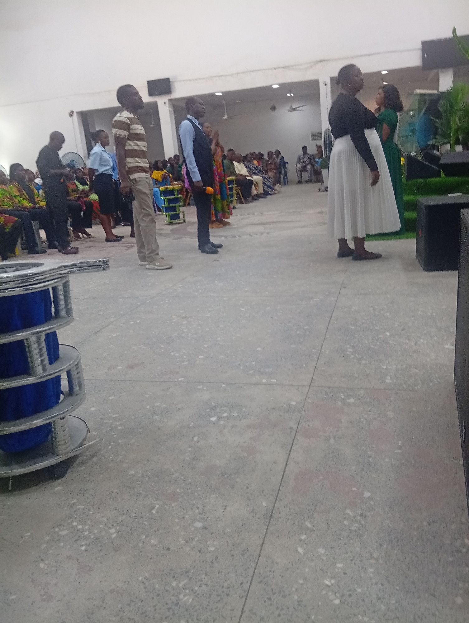 In Kason, Nigeria, a group of people stand in what appears to be a church or event hall, facing towards the front of the room. In the foreground, to the left, is a stack of large industrial spools, likely holding cables, with blue material wound around them. The floor is a light-colored, speckled concrete.
Further back, a man in a striped shirt and khaki pants stands with his head tilted upwards. Beside him, a man in a blue shirt and vest holds a microphone. To the right of them, two women are positioned. One wears a black long-sleeved top and a flowing white skirt, and the other is in a green dress.
In the background, other attendees are seated or standing, many wearing colorful attire. The room is brightly lit with overhead lights, and a ceiling fan is visible. The overall atmosphere seems to be one of a gathering, possibly a religious service or a formal presentation.