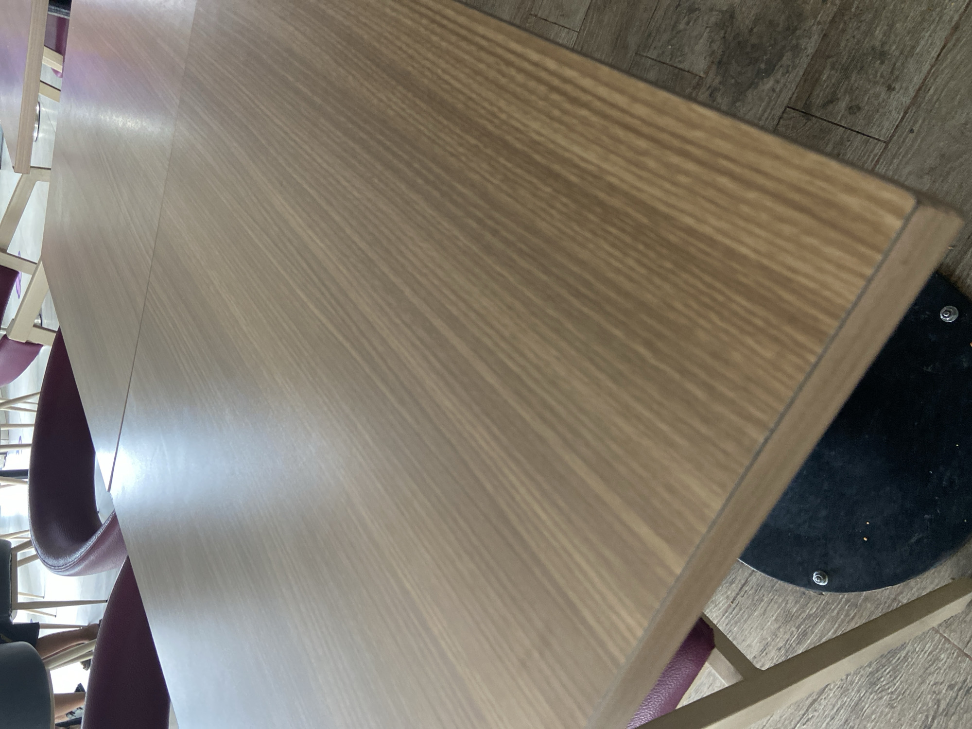 This is a close-up, eye-level shot of a light brown, wooden table with a visible grain pattern. The table appears to be in a public space, possibly a cafe or restaurant, as indicated by the presence of purple chairs in the background. The floor is made of grey, wood-look tiles arranged in a herringbone pattern. The table has a dark, circular base with a visible screw. The scene is indoors, with soft lighting creating reflections on the table's surface. There are no people clearly visible in the foreground or the main focus of the image. The image is devoid of any identifiable landmarks or context that would place it specifically in Bago, Indonesia.