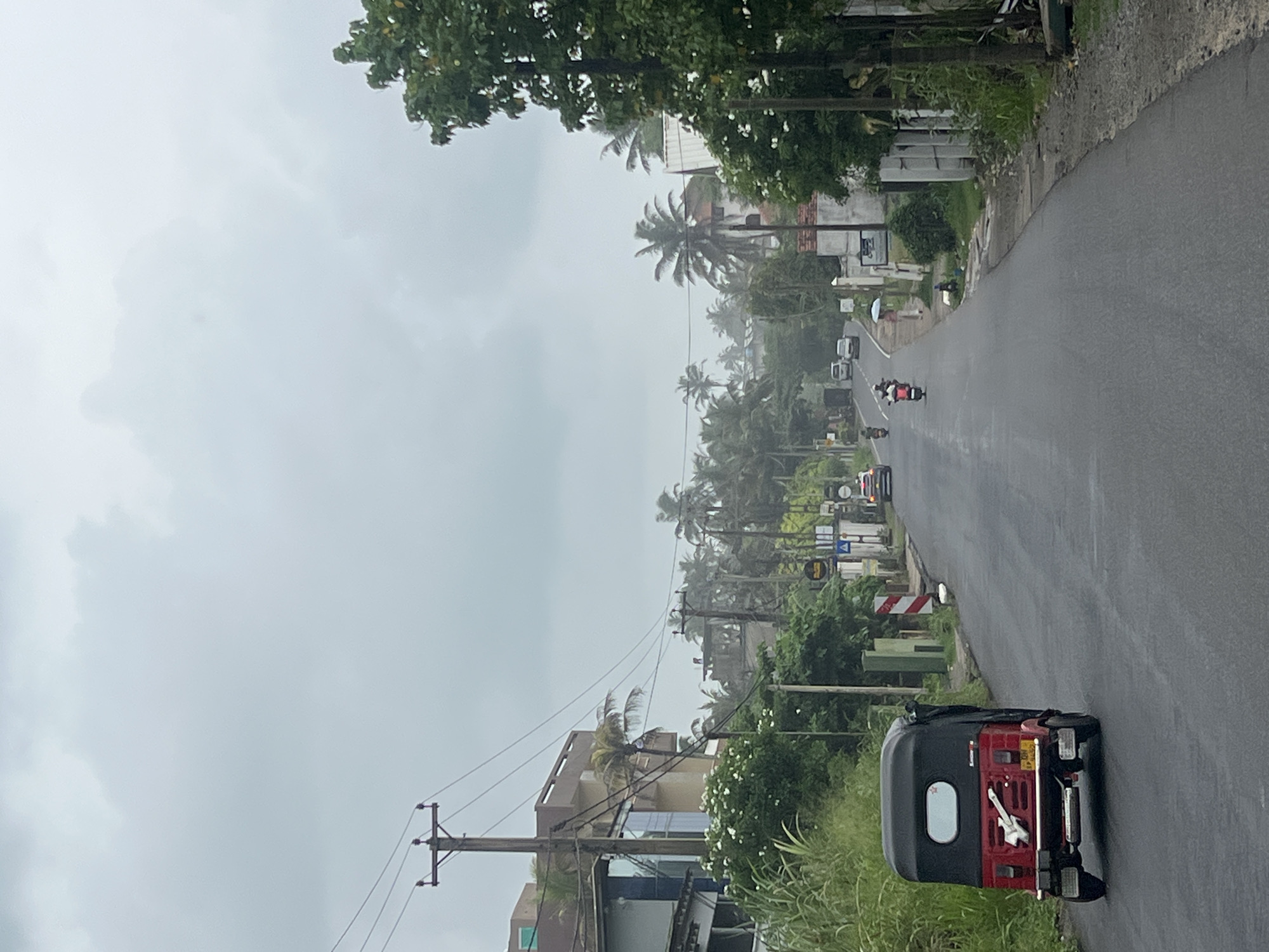 A road in Goviyapana, Sri Lanka, stretches into the distance, lined with palm trees and buildings under a cloudy sky. In the foreground, a three-wheeled auto-rickshaw sits parked on the right side of the road. Further down the road, a few cars and motorbikes are visible. Power lines are strung along the roadside.