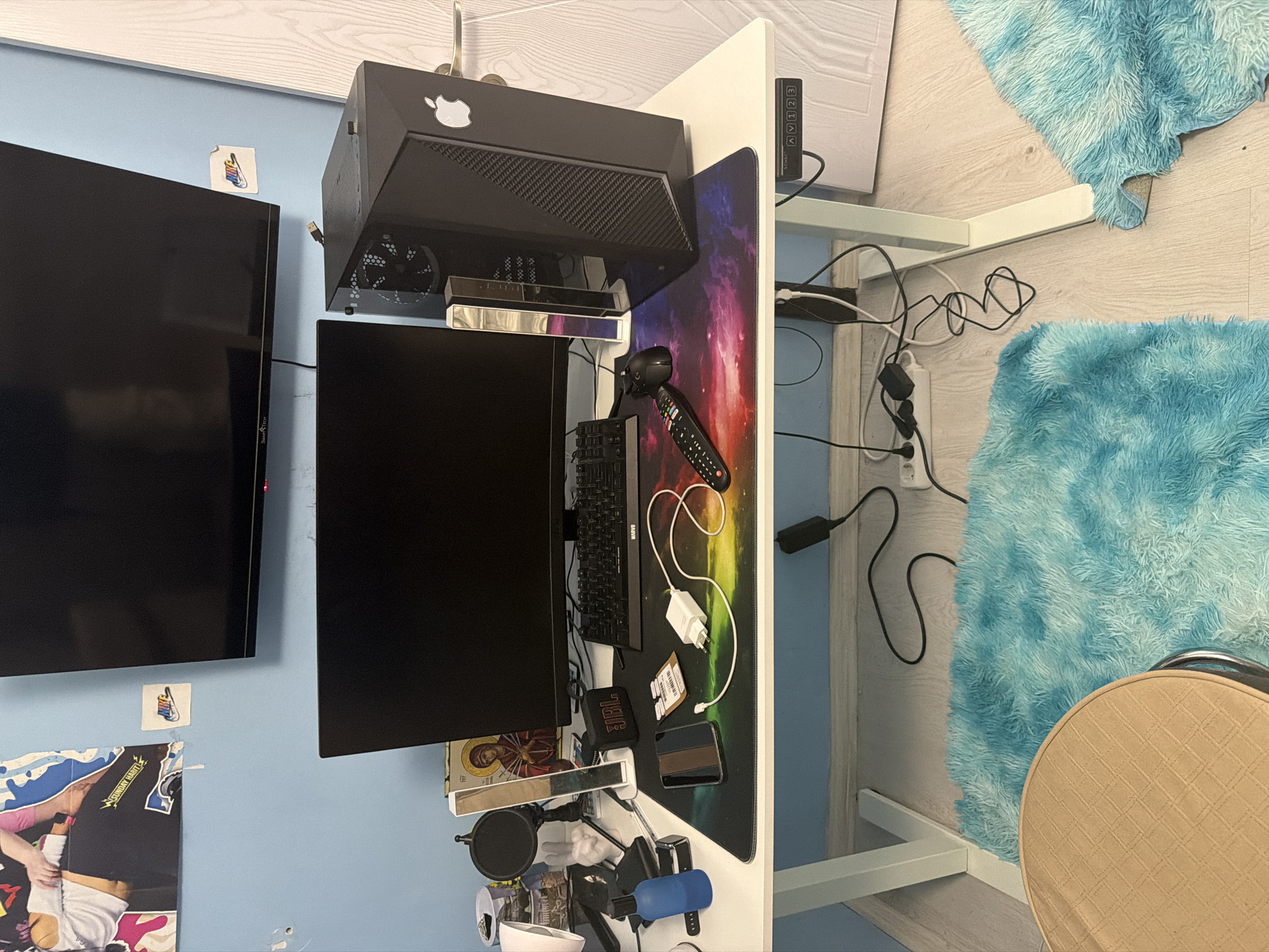 The image captures a personal computer and entertainment setup within a room, viewed from an overhead perspective, with the original image oriented vertically (rotated 90 degrees clockwise from natural orientation). A white desk, likely positioned against a light blue wall, dominates the central area.
On the desk, a black desktop computer tower with an Apple logo sticker sits on the left side, above a black computer monitor. Below this monitor, a silver soundbar or similar device is visible, along with a microphone and pop filter. A black keyboard and mouse are positioned on a large gaming mouse pad featuring a colorful galaxy design. To the right on the mouse pad are a black universal remote control, various charging cables, a white power adapter, a JBL portable speaker, and a black wallet or cardholder. A small blue bottle is also on the desk.
To the left of the desk, mounted on the light blue wall, is a large black flat-screen television or monitor with "SmartTech" subtly visible on its side. Below it, a poster with a partial image of a person wearing a t-shirt that reads "SUNDAY HARDT" is visible, along with an icon or religious image. Above the larger monitor, a small paper with a Nike logo is stuck to the wall. Another small paper with a colorful design is visible below the computer tower.
To the right of the desk setup, against a white dividing panel, a multi-input selector box with "AV 1 2 3 TV" printed on it is mounted. The floor is made of light-colored wood, covered in parts by two fluffy, light blue rugs. A beige, round chair with a textured, checkered pattern is partially visible in the bottom right corner. Various power cables and extension cords are visible on the floor beneath the desk, connecting to power outlets. The room appears to be a personalized space, possibly a bedroom or home office.