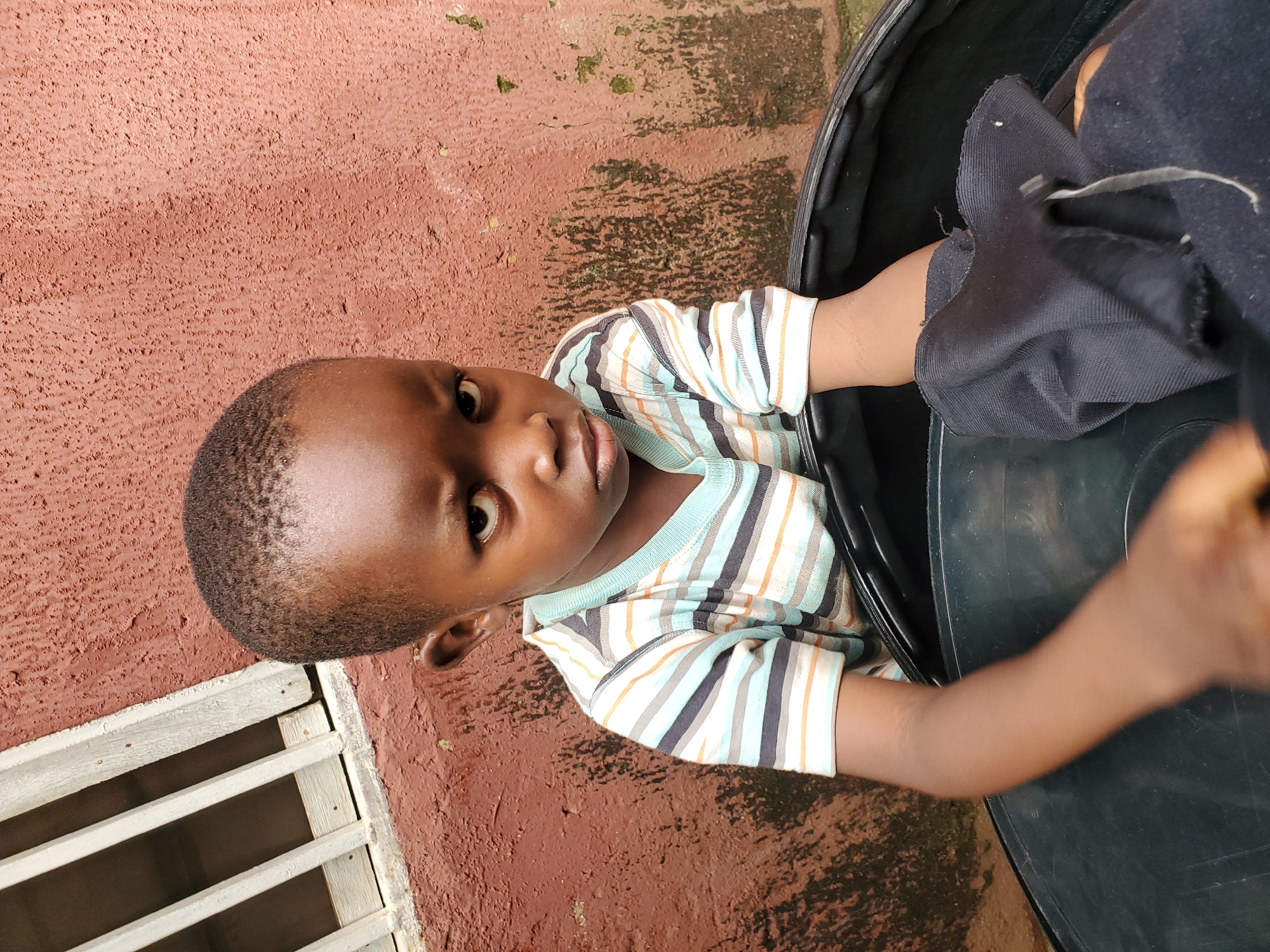 A young child, appearing to be a boy, is captured from a high angle in what appears to be an outdoor setting. He is wearing a striped t-shirt with horizontal lines of white, blue, orange, and gray. The child is situated within or next to a large, black object, possibly a container or a drum. His arms are visible, with his right arm extending towards the black object.
The background consists of a textured, reddish-brown wall, which shows signs of wear and patches of green algae or moss, suggesting an outdoor environment that may experience rain or humidity. To the left of the child, a section of a white wooden structure with parallel slats is visible. This structure could be a fence or a window grate.
The child's gaze is directed towards the camera with a slightly curious or contemplative expression. The lighting suggests it is daytime, though the specific time is not discernible. The overall impression is a candid moment, possibly in a residential or communal area in Jos, Nigeria, given the provided location context. No visible text is present in the image.