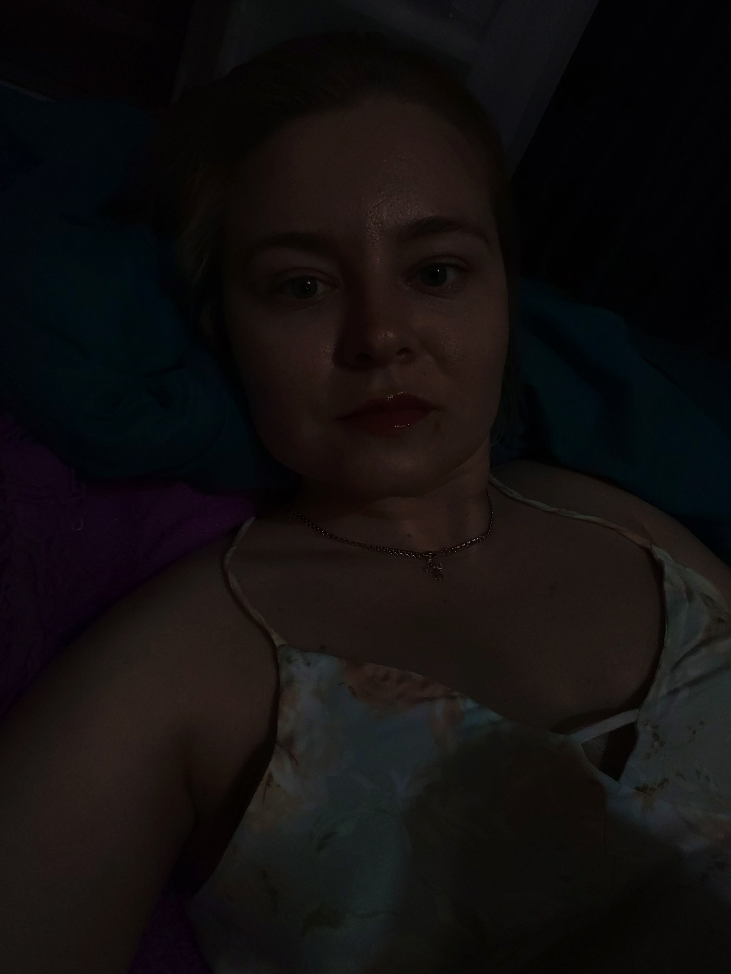 A woman is reclined, possibly on a bed, in a low-lit environment. She is wearing a light-colored tank top with floral patterns and a silver chain necklace. The background consists of a dark blue pillow and what appears to be a purple fuzzy surface. The location in Izhevsk, Russia is not discernible from the image.