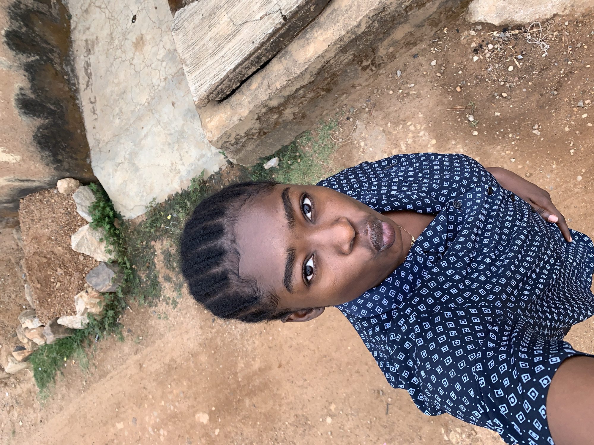 A medium-angle, eye-level shot depicts a dark-skinned person in Rafin Par, Nigeria. The individual is wearing a dark blue button-down shirt with a small white geometric pattern. Their hair is styled in neat, short braids. The person is positioned on a dirt ground with sparse vegetation. Concrete stairs and a retaining wall are visible on the left side of the frame. The overall lighting is bright and natural.