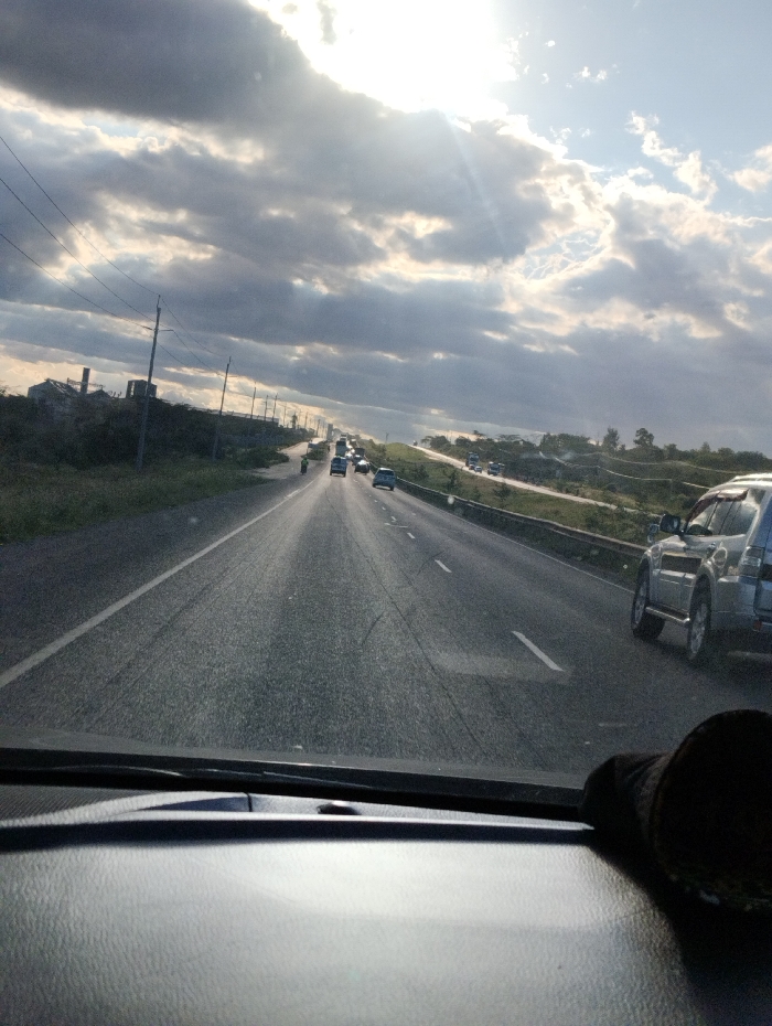 The image shows a view from inside a vehicle traveling on a highway in Athi River, Kenya, at coordinates -1.497 latitude and 37.074 longitude. The highway has a few cars traveling in both directions. The sky is bright with sun shining through the clouds. The vegetation is sparse and the landscape is flat.