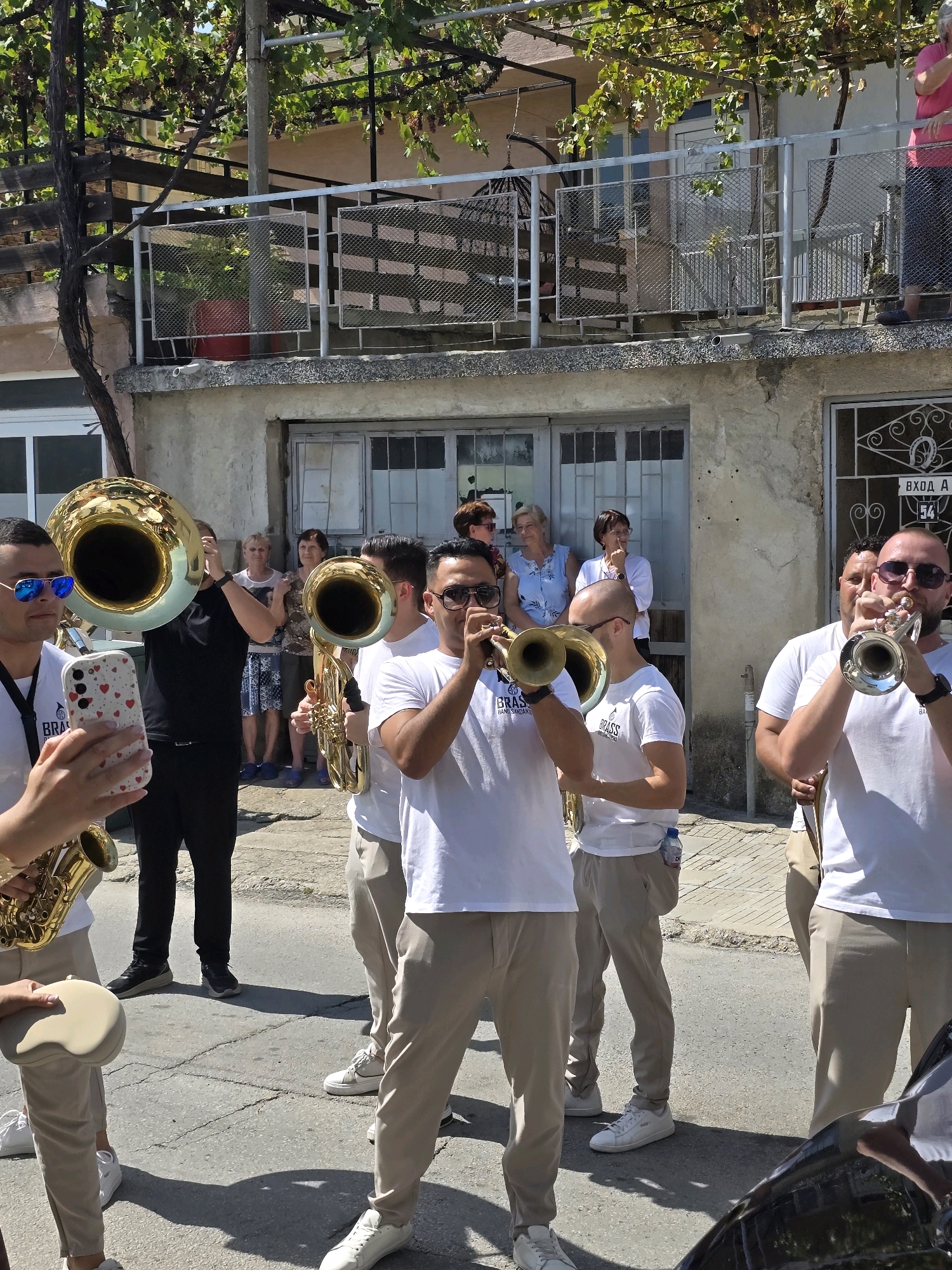In Polenitsa, Bulgaria, a brass band performs outdoors on a street. The band members, all male and wearing white t-shirts and tan trousers, play various brass instruments including tubas, a saxophone, and trumpets. The foreground features a man in sunglasses holding a smartphone and a saxophone. Behind him, another band member plays a tuba. In the background, several women observe the performance. The setting includes a building with an upper balcony and grapevines overhead, suggesting a warm, sunny day. The street appears to be paved, and shadows are cast on the ground.
