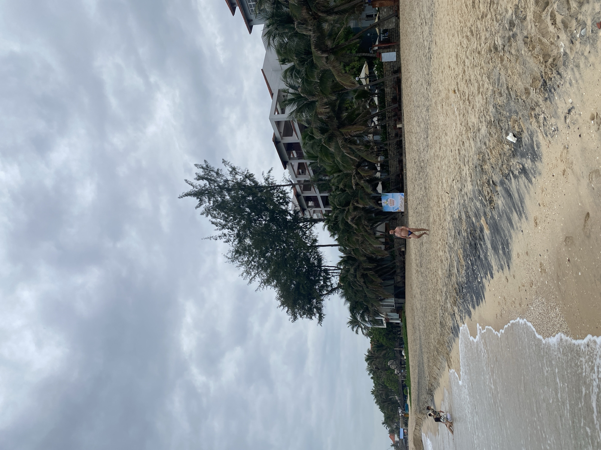 The image depicts a beach scene in Phu Thuy Ward, Vietnam, under a cloudy sky. A multi-story building stands adjacent to the beach, separated by a line of palm trees and other greenery. The beach is sandy, with a darker band running parallel to the shoreline. The waves are gentle, and at least three people are visible: one standing alone on the sand closer to the building and two more together walking in the water closer to the bottom of the image. The vegetation includes palm trees and what appears to be an evergreen-type tree. A sign stands near the palm trees.