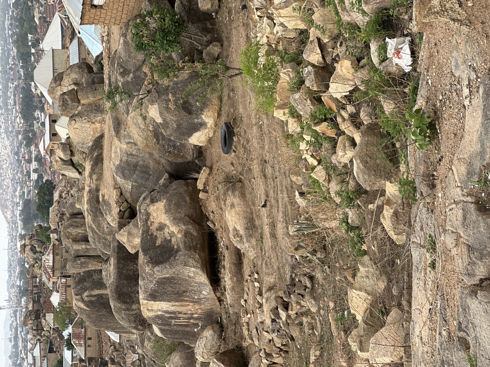 The image depicts a rocky landscape, seemingly a hillside, in the city of Jos, Nigeria. Large, rounded rock formations cascade down the slope, interspersed with patches of dry earth and sparse vegetation. A discarded car tire sits in the middle of the terrain. Scattered rocks and debris are present, as well as a white piece of trash. Below, the skyline of Jos is visible, characterized by dense clusters of buildings with corrugated iron roofs. The environment appears arid and somewhat rugged, suggesting a semi-urban landscape on the outskirts of the city.