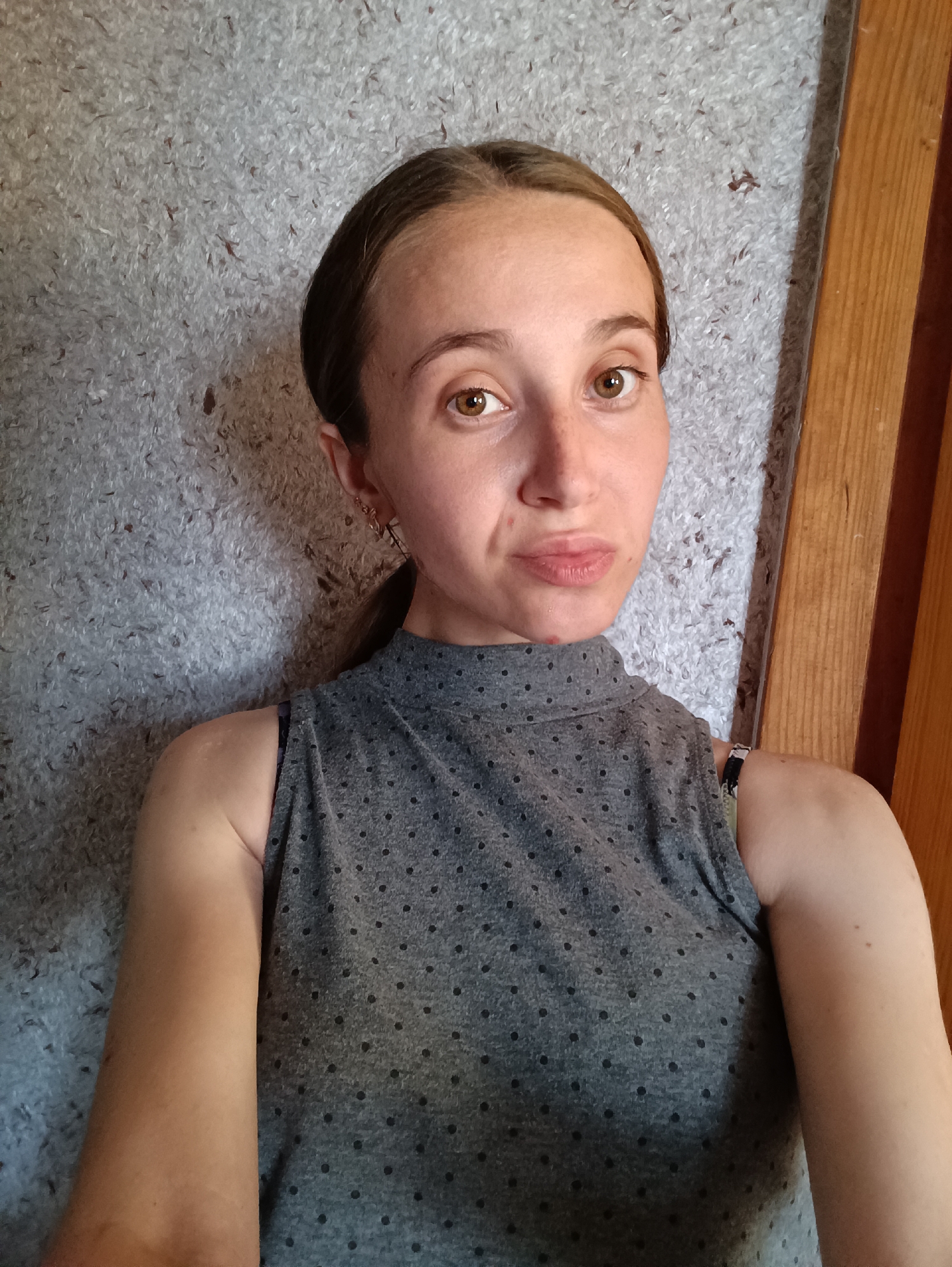 A woman is looking at the camera in a selfie. She has light skin, brown eyes, and her dark brown hair is pulled back in a ponytail. She is wearing a grey sleeveless top with a black polka dot pattern and a black bra strap is visible. She is also wearing a delicate gold earring. The background appears to be a wall with a textured, grey surface, possibly a textured wallpaper, and to the right, a vertical wooden beam. The scene is indoors and the lighting is natural, suggesting daytime. There is no discernible activity or interaction. The image does not provide any information about the location within Vypasne, Ukraine.