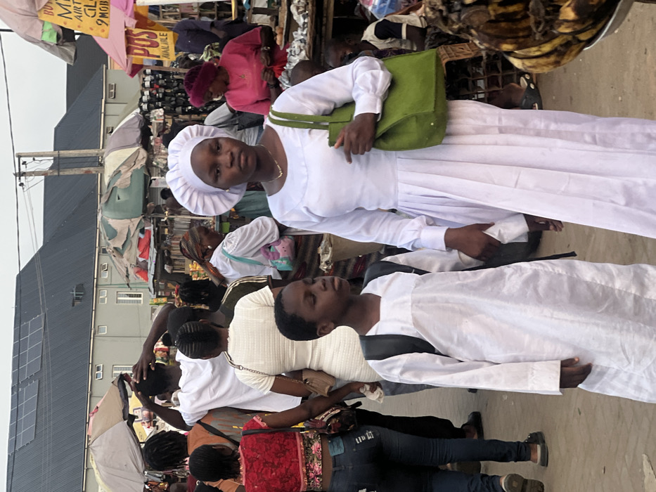 A group of people are walking through an outdoor market. Two people dressed in white robes and hats are prominent in the foreground. The woman has a green purse on her shoulder. The market is bustling with people and vendors. Merchandise and signs are visible among the crowd. The scene appears to be in Nigeria based on clothing and building architecture.