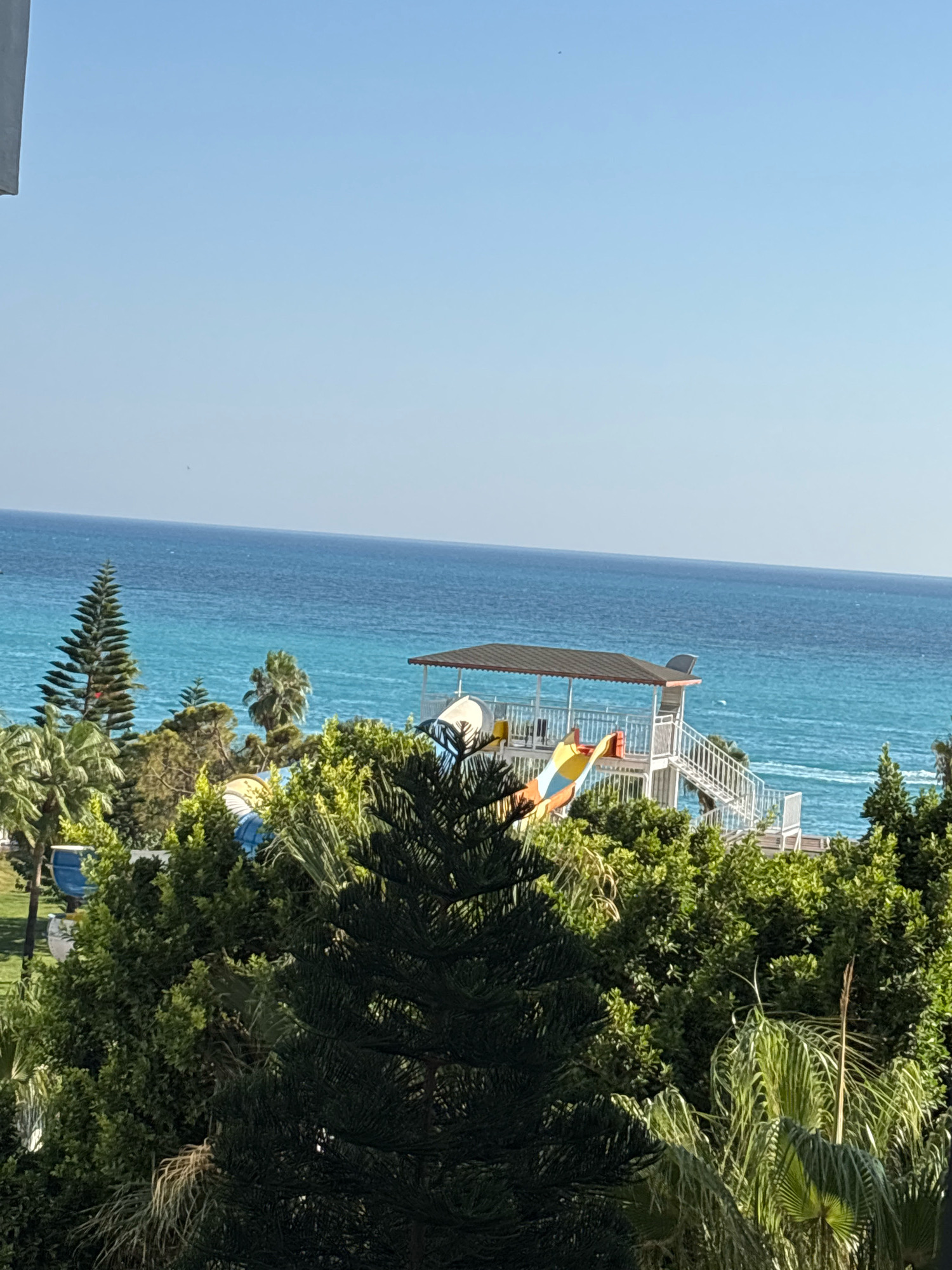 This is an outdoor, eye-level, horizontal, full shot of a resort area with a view of the Mediterranean Sea. The sky is clear and blue. In the foreground, there are various types of green trees, including a tall, dark green conifer tree on the left. In the middle ground, there is a white structure with a brown roof, which appears to be part of a water slide. The slide itself is colorful with yellow, red, and blue sections. White stairs with railings lead up to the structure. Behind the water slide and trees, the bright turquoise Mediterranean Sea stretches to the horizon. The water shows some white ripples and waves. To the left of the water slide structure, a glimpse of blue is visible, possibly indicating a swimming pool. The overall impression is a sunny day at a tropical resort, likely in Alanya, Turkey, given the characteristic blue sea and lush vegetation. There are no people visible in the image.